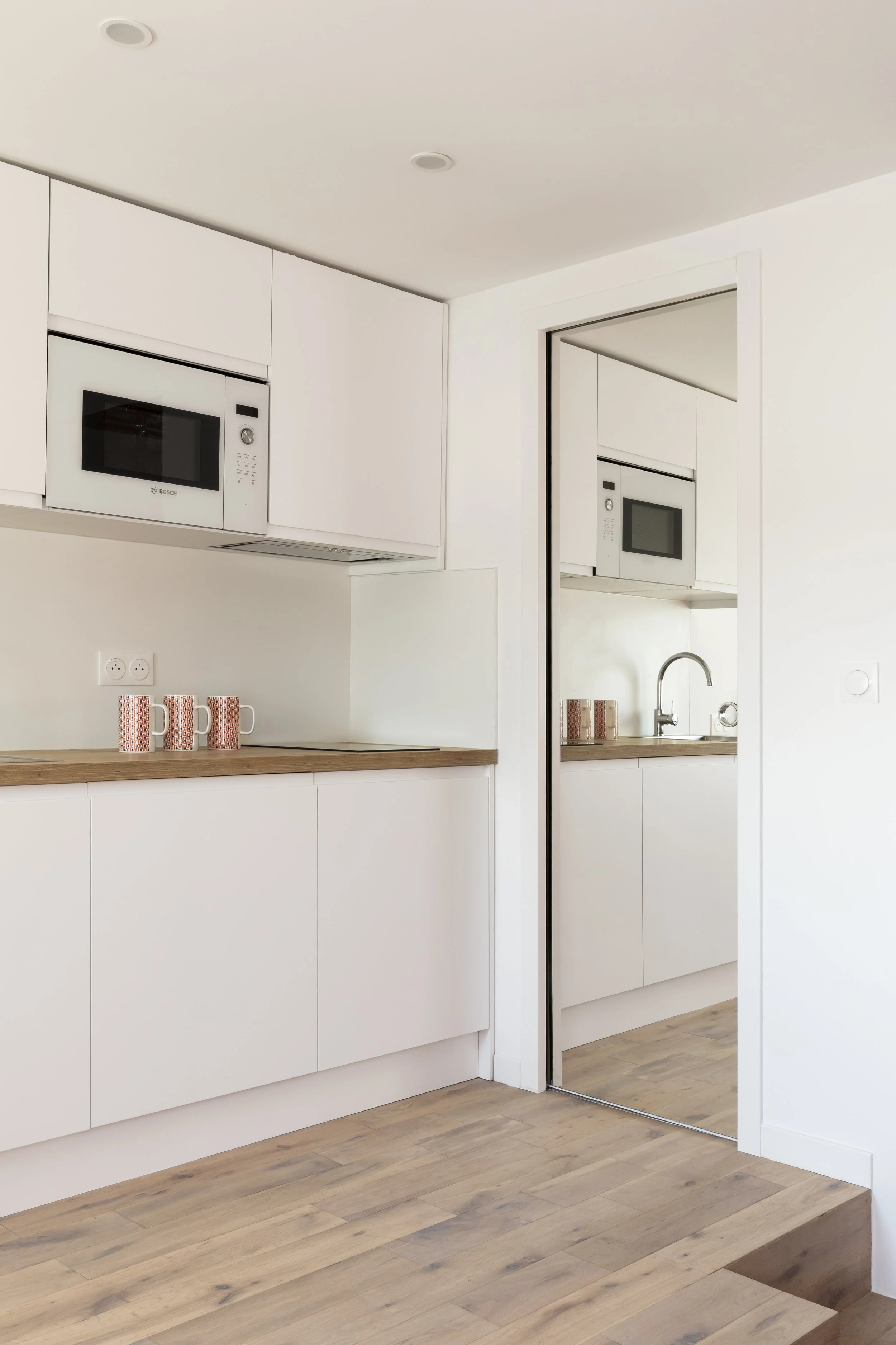 Cuisine moderne avec des armoires blanches, un micro-ondes intégré, un plan de travail en bois, un évier avec robinet, et un miroir servant de porte à une autre pièce.