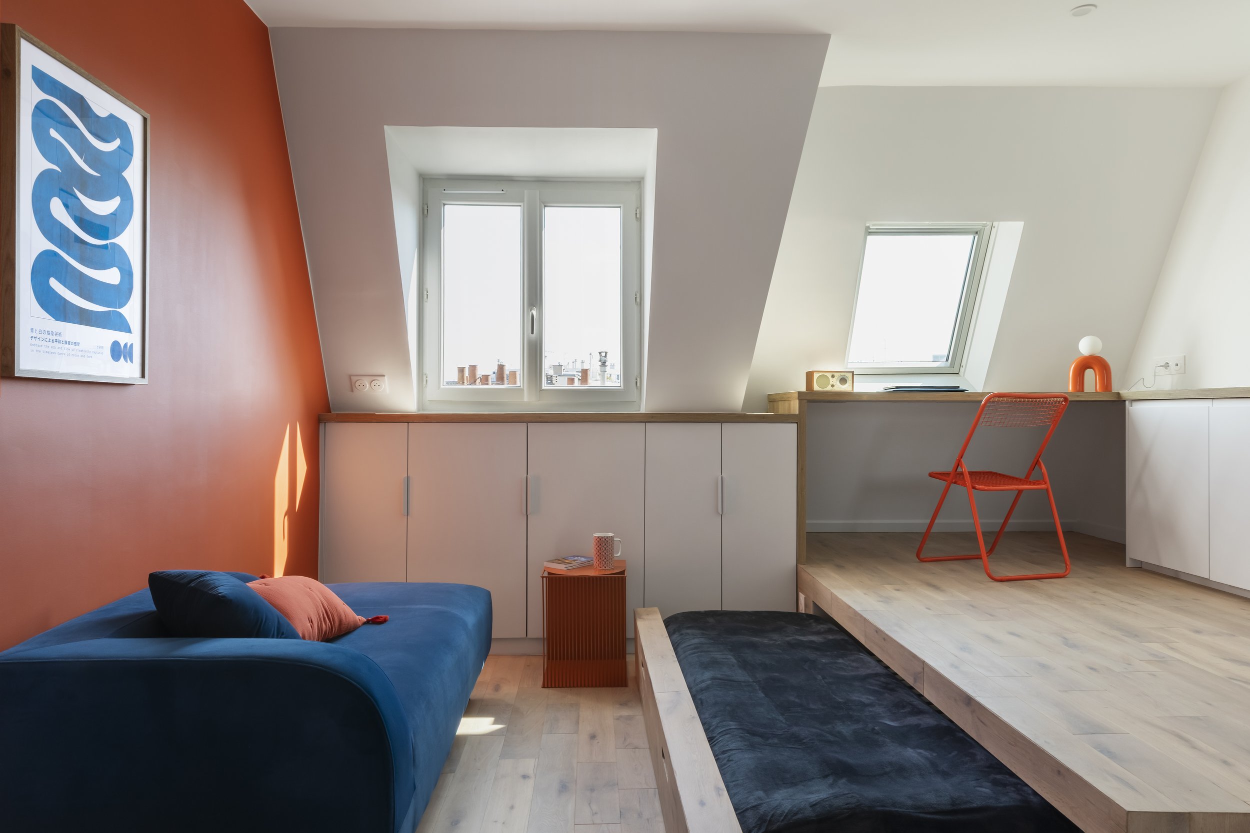 Intérieur d’un loft avec murs blanc et orange, deux fenêtres, un canapé bleu avec coussins orange, une petite table orange avec une tasse et des magazines, un escalier en bois, un fauteuil orange, une lampe orange et une petite Radio sur une étagère.