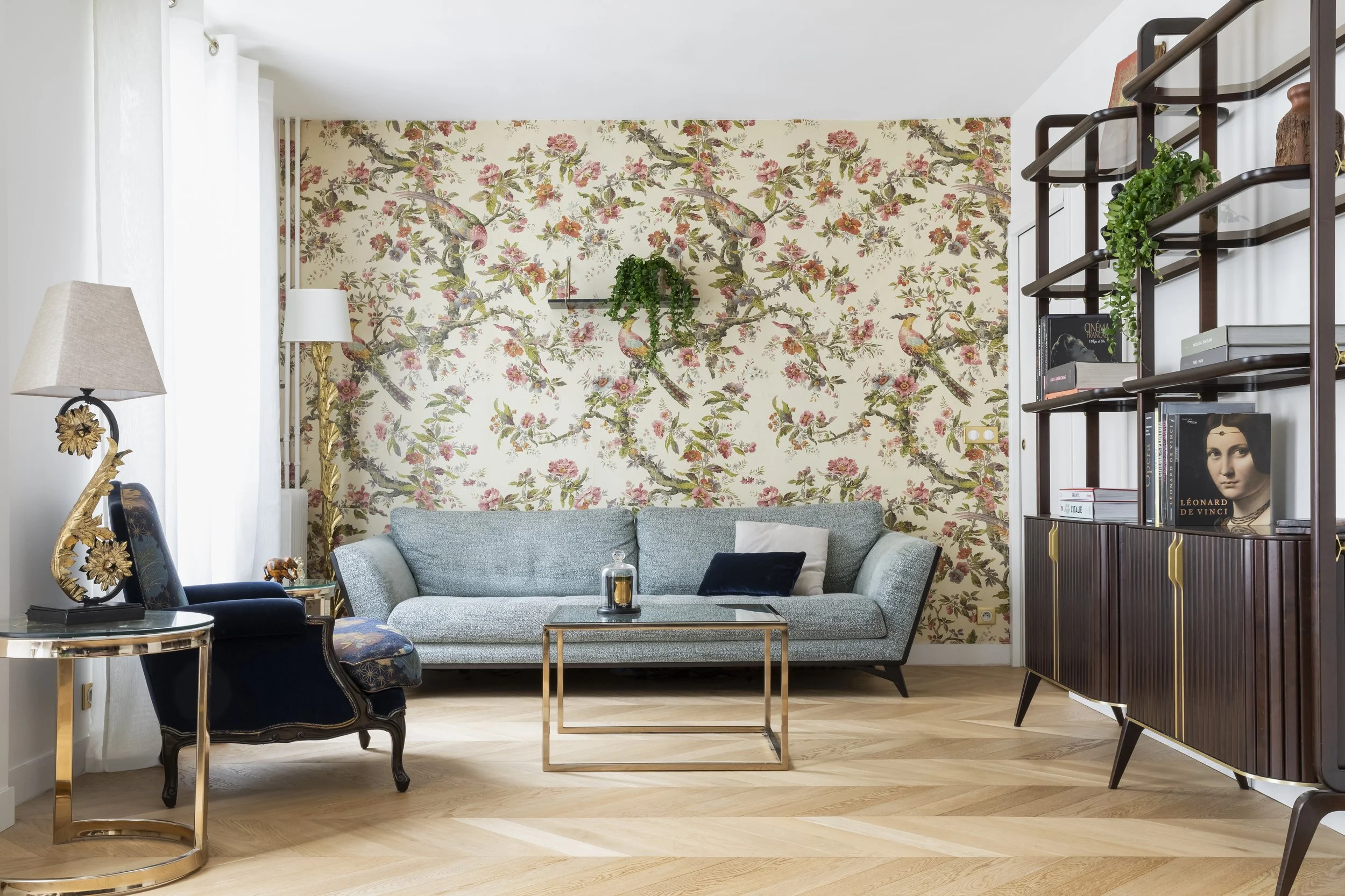 Salon avec un mur à papier peint floral, canapé gris clair avec coussins blancs et noirs, table en verre avec cadre en or, étagère en bois avec livres et décoration, lampe avec abat-jour beige, chaise en velours bleu foncé avec détails dorés, sol en 
