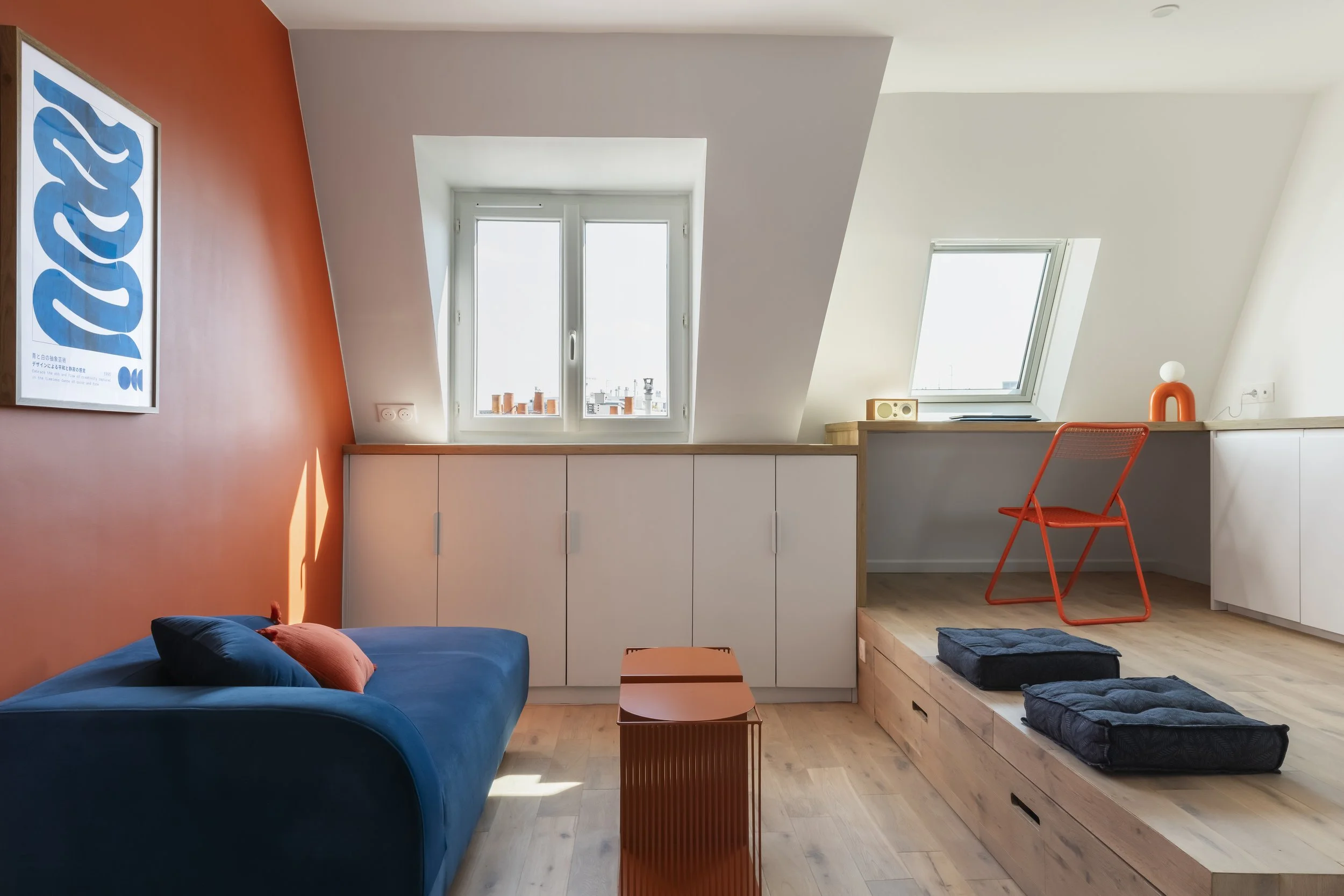 Intérieur d'un loft avec deux fenêtres, un mur orange avec une affiche bleue, un canapé bleu avec des coussins, un meuble blanc, une chaise orange, un lit en bois avec des literies grises, et un plafonnier blanc.