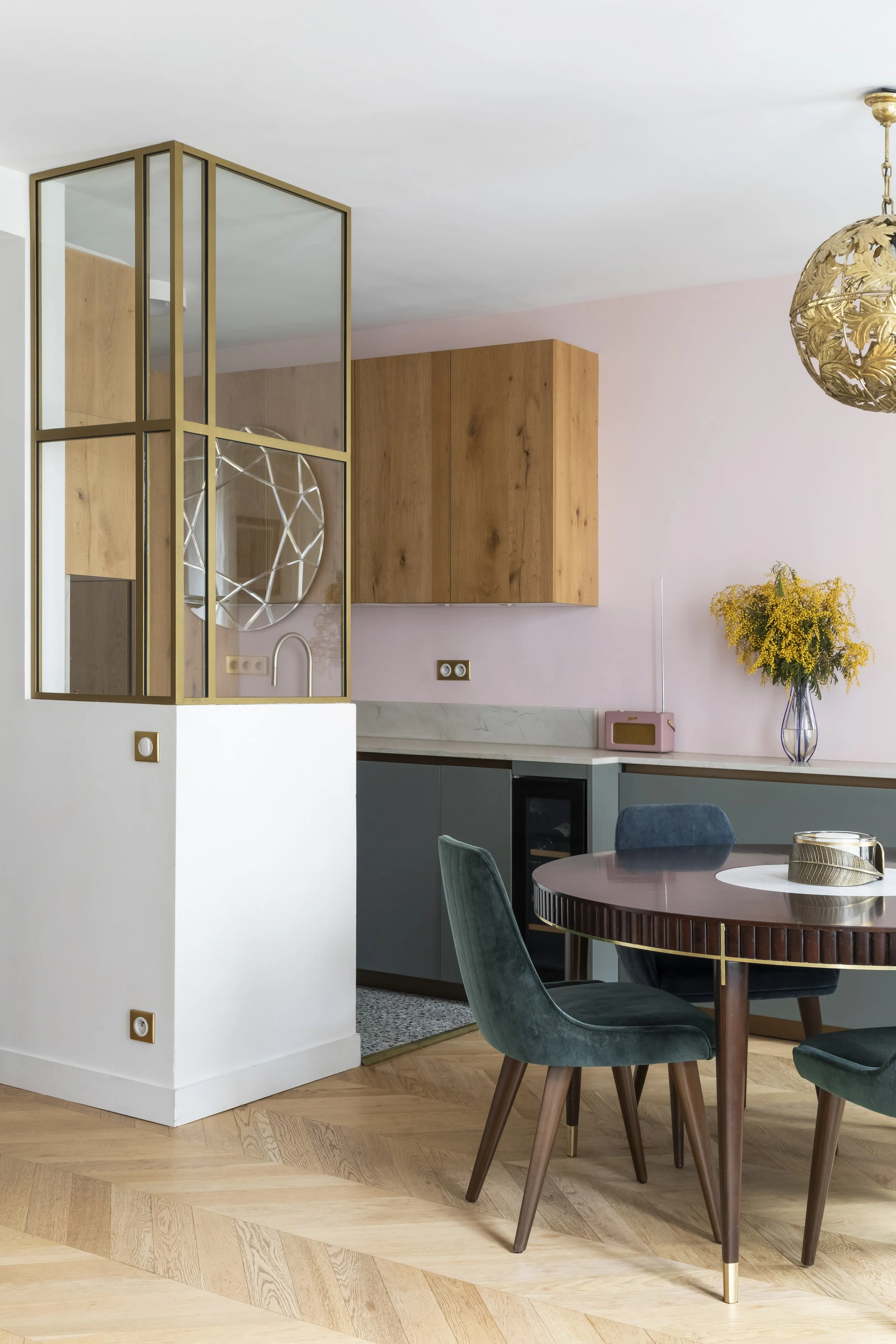 Intérieur moderne d'une cuisine et salle à manger avec un mur rose clair, des meubles en bois, une table ronde en bois foncé avec des chaises vert foncé, une lampe en métal doré, un vase avec des fleurs jaunes, un four intégré et un radiateur rose.