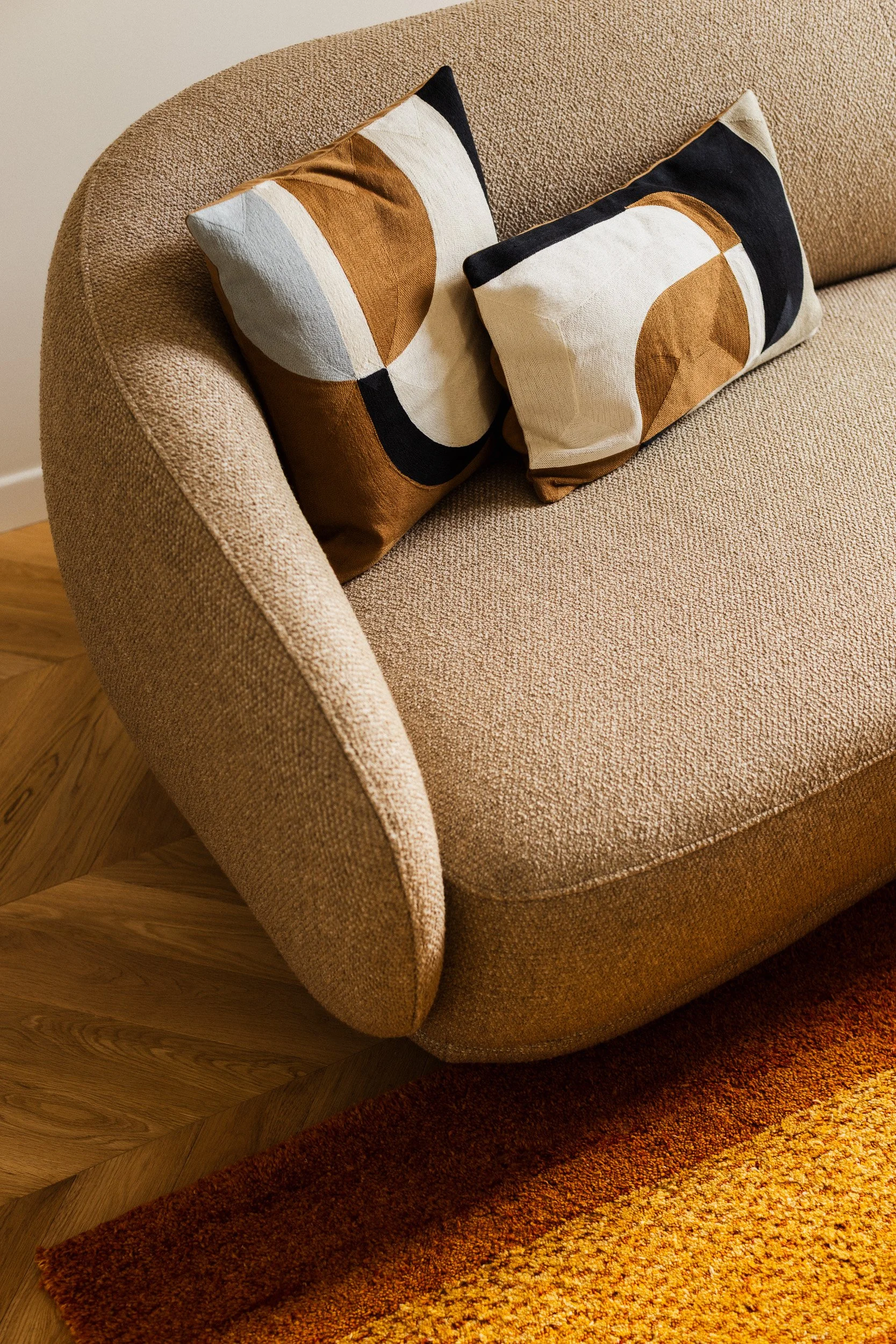 Un canapé beige avec deux coussins géométriques de couleurs marron, noir, blanc et beige. Le canapé de la marque Boconcept modèle Bolsano est placé sur un parquet en bois clair en point de Hongrie, avec un tapis aux couleurs chaudes au sol.