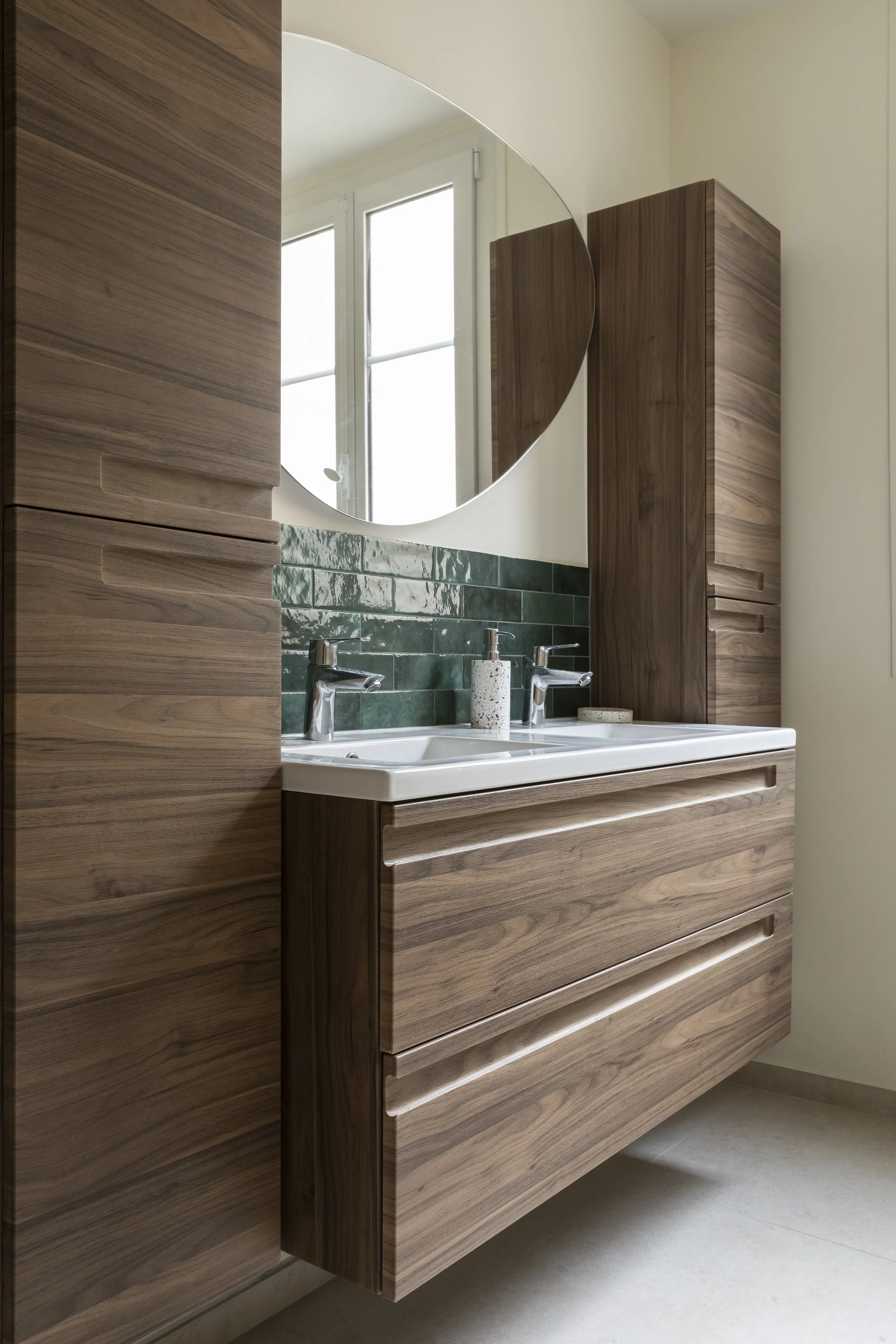 Un meuble de salle de bain en bois avec deux tiroirs, un miroir rond au-dessus, un lavabo double, un savon en pompe blanc et un chiffon.