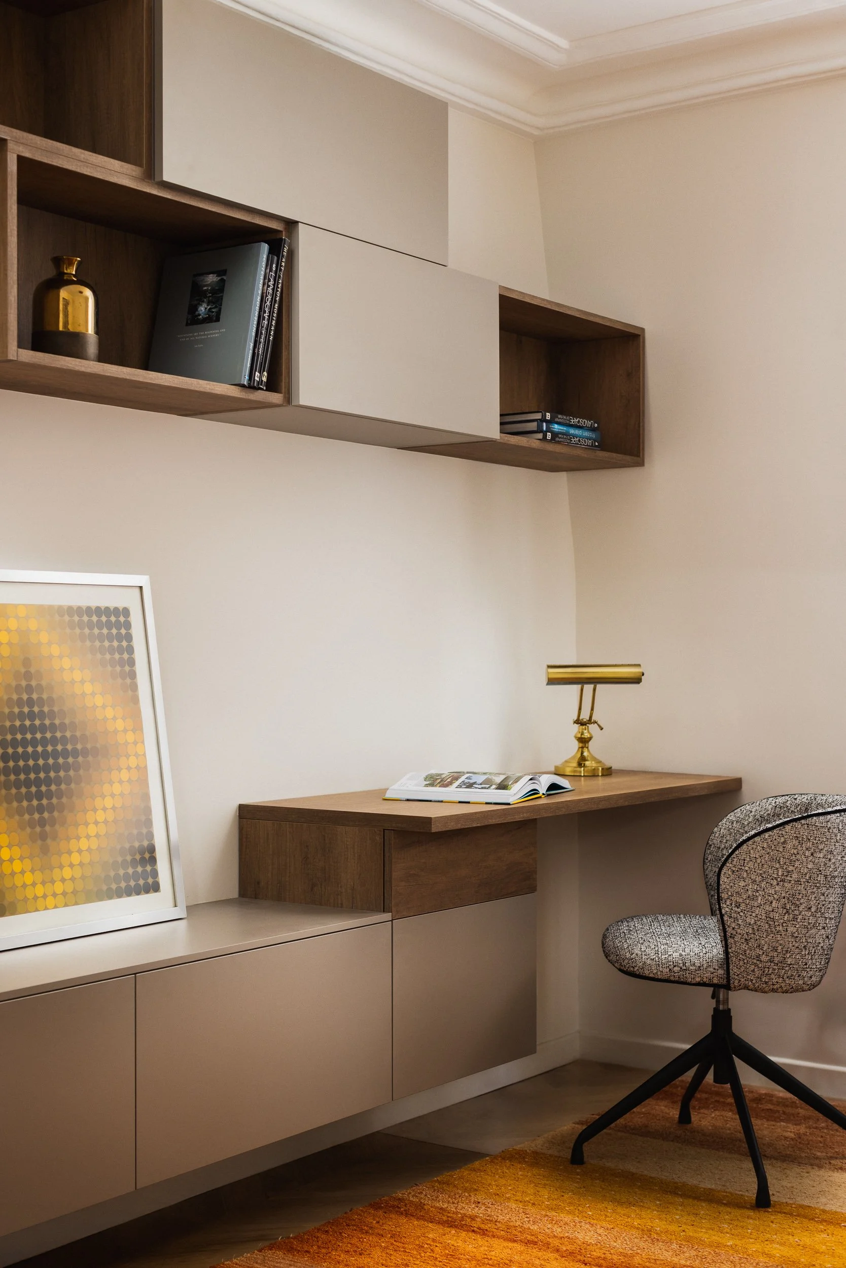 Un espace de bureau minimaliste dans un intérieur Haussmannien avec un mur blanc, un bureau suspendus et des rangements niches et placards suspendus en bois, étagères en bois , une lampe de bureau doré, une chaise de bureau en tissu à motifs, et un t