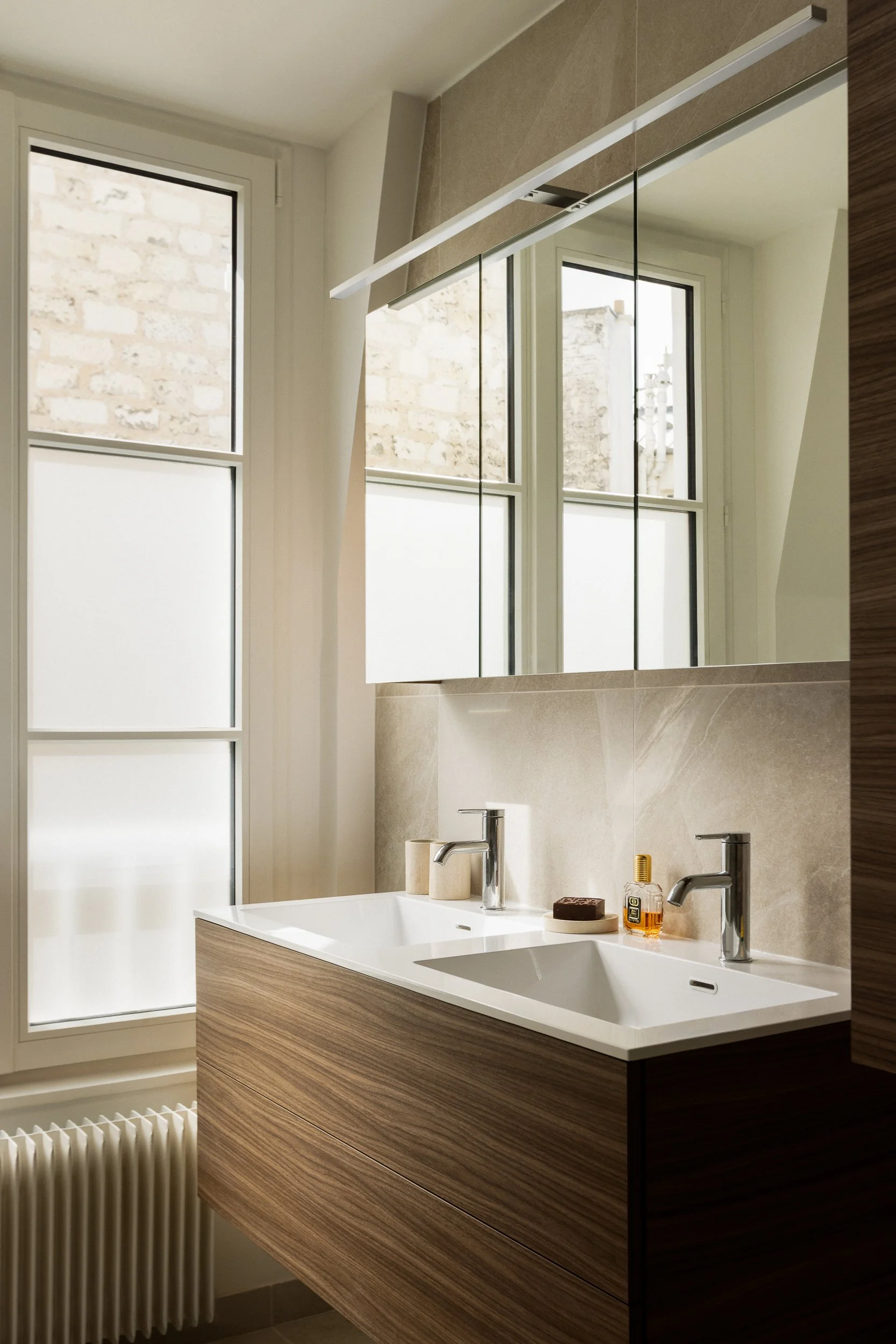 Vue d'une salle de bain moderne avec deux lavabos, un grand miroir, fenêtres avec vue sur un mur en pierre, et éléments de décoration en beige et bois.