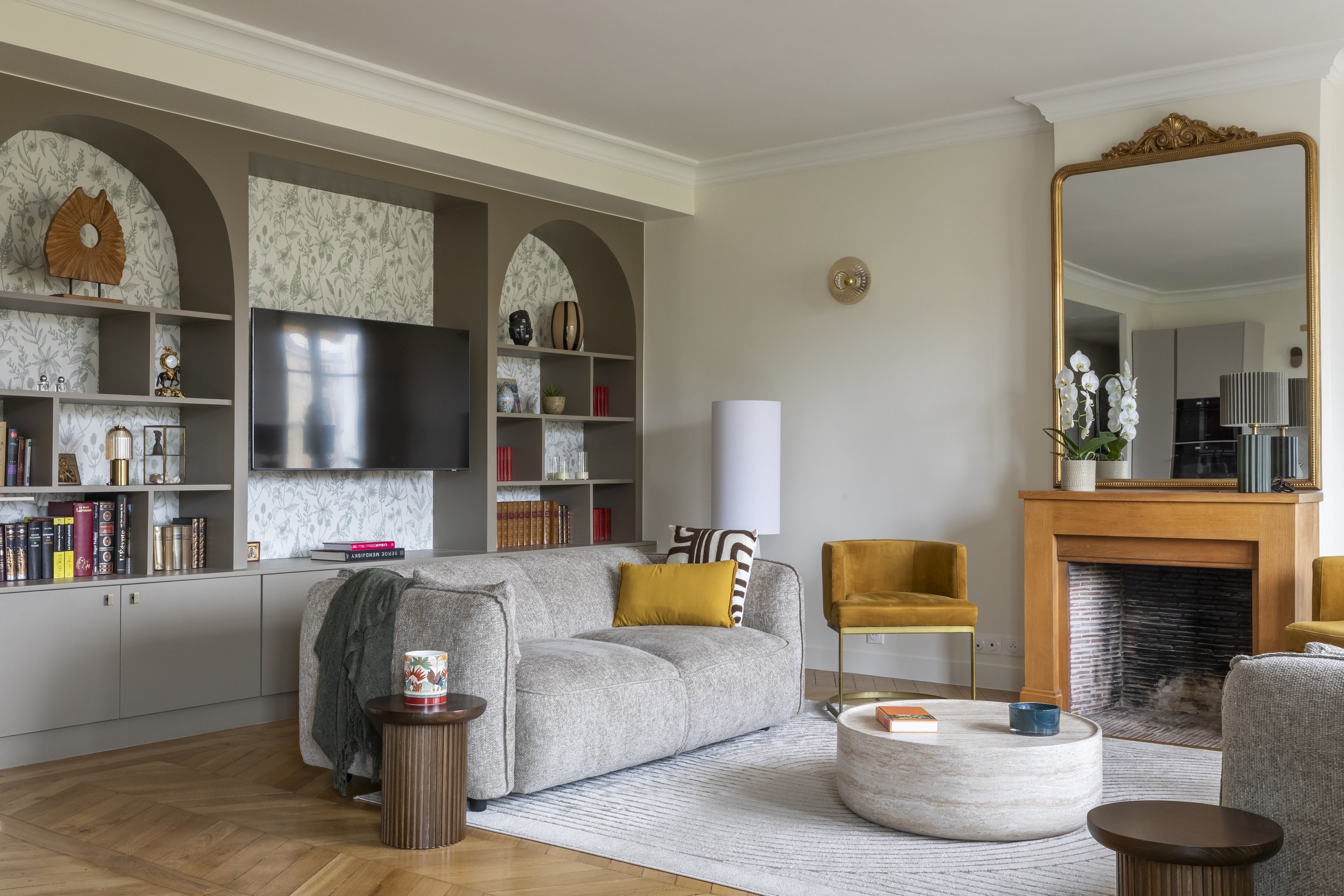 Un salon moderne avec canapé gris, tapis blanc, tables basses en bois, cheminée en briques, grande miroir encadré doré, étagères avec décorations, télévision fixée au mur, lampes, et une plante en pot avec des fleurs blanches.