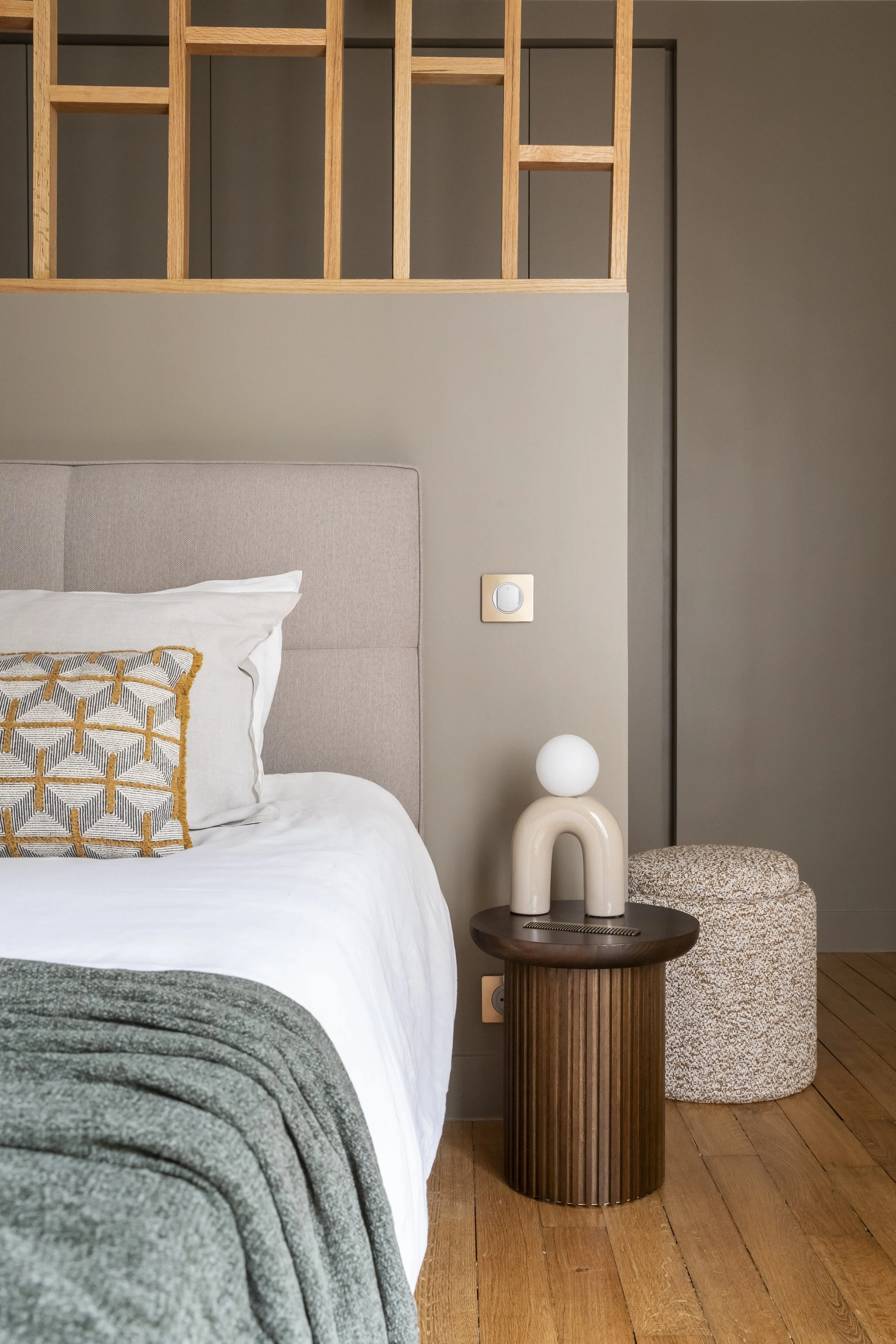 Coin de chambre avec un lit, une table de nuit en bois, une lampe décorative, un fauteuil rembourré, et un mur avec des détails en bois.