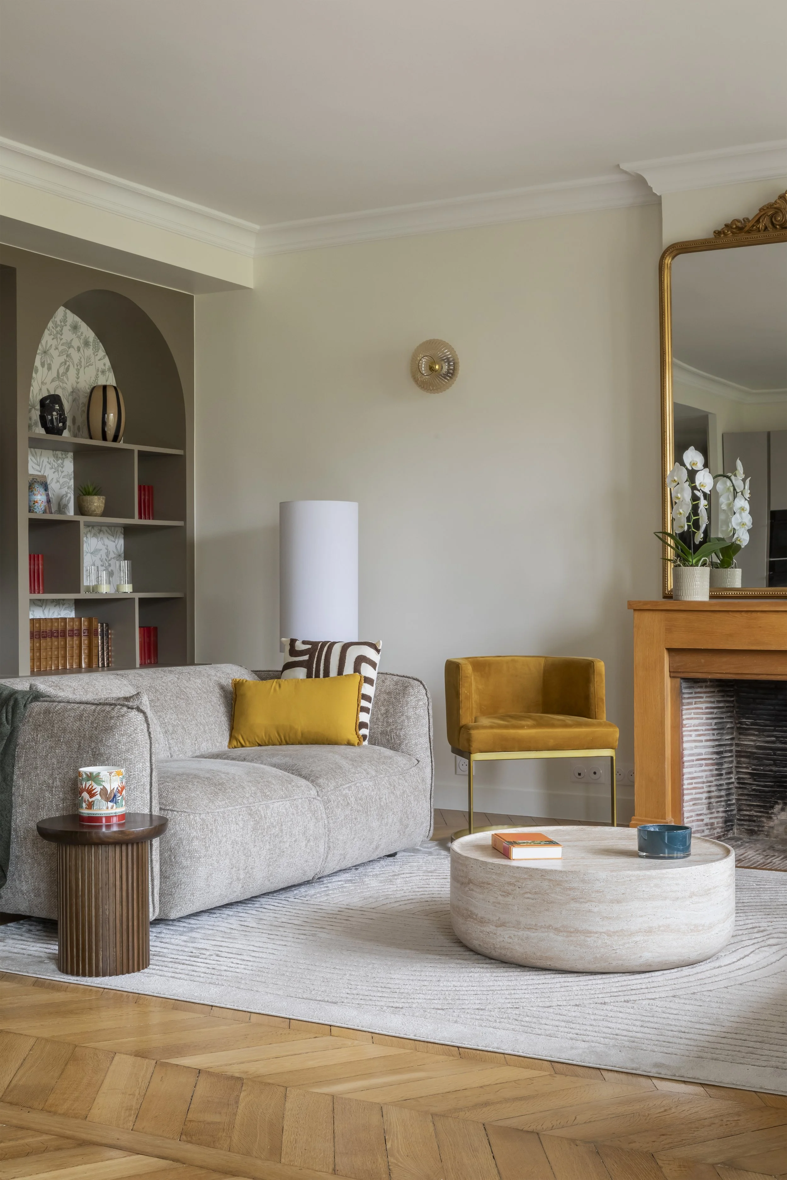 Salon moderne avec canapé beige, table basse ronde en pierre, chaise jaune, cheminée en bois avec miroir doré, étagère avec livres et décorations, lampadaire blanc, plantes tropicales en pot, tapis côtelé beige, parquet en bois.