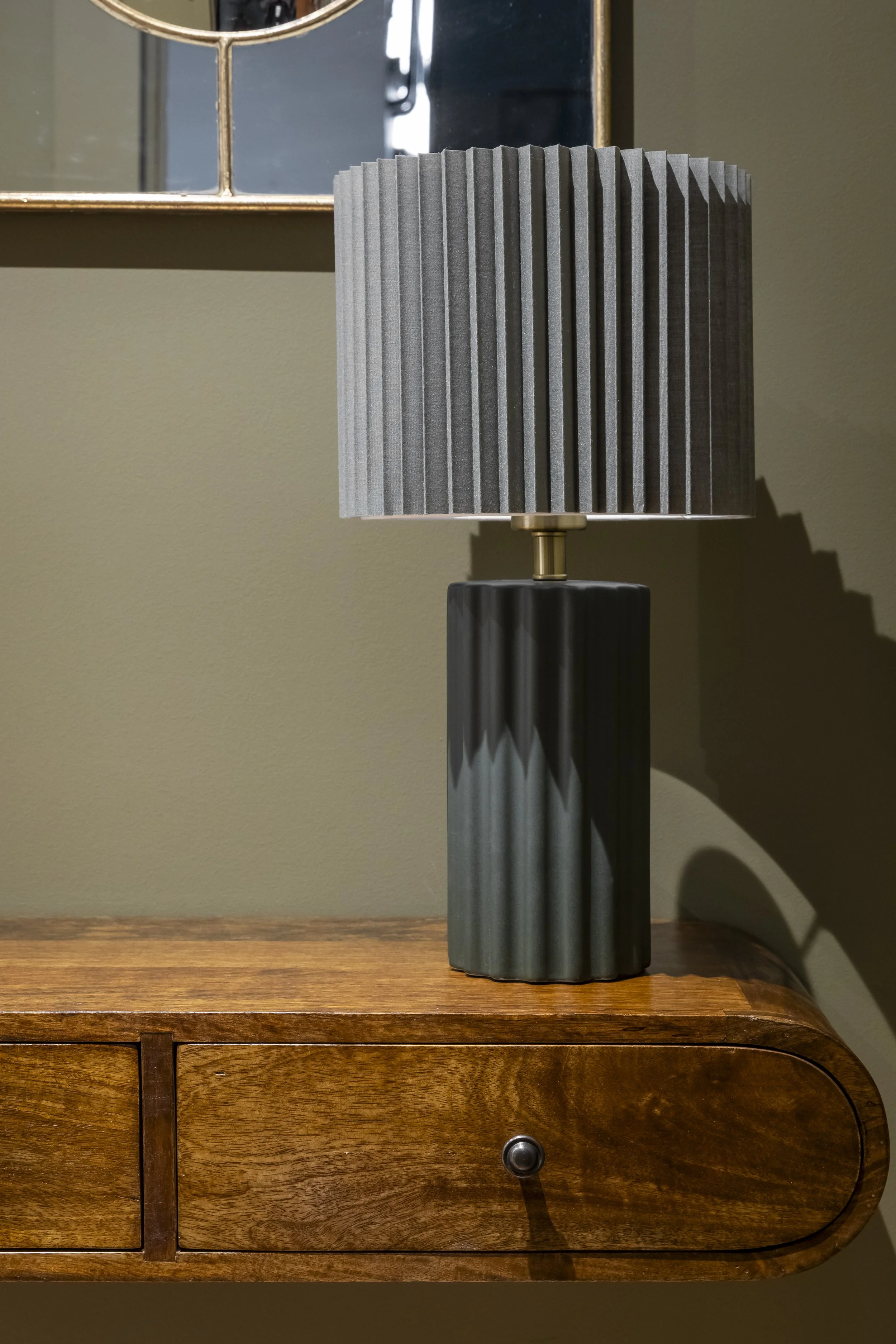 Une lampe de table moderne avec un abat-jour en tissu gris à pli vertical, placée sur une surface en bois massif. Un miroir carré à cadre métallique doré est visible en arrière-plan.