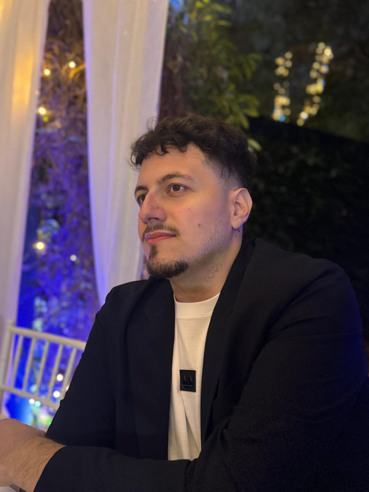 Uomo seduto in un ambiente all'aperto di sera, con luci soffuse e decorazioni luminose sullo sfondo.