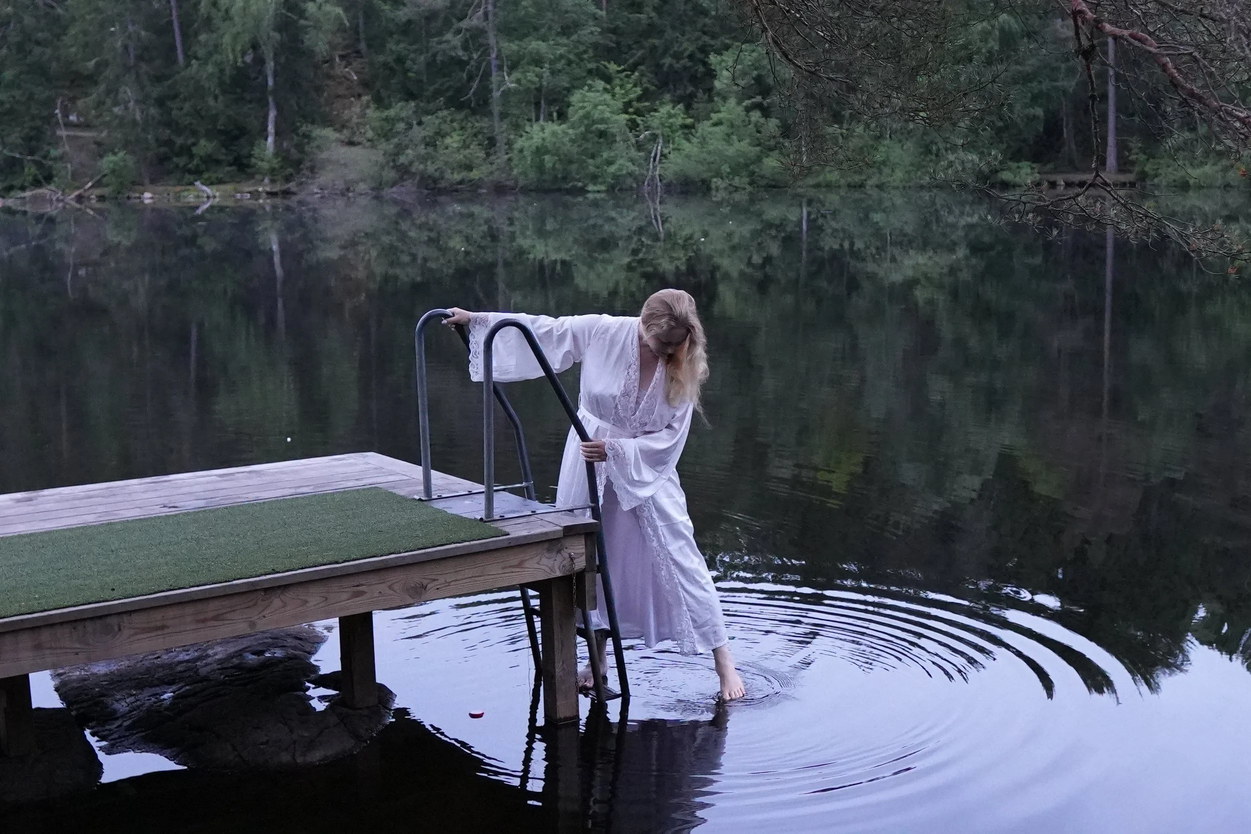 A woman in a white dress with lace details is standing on a dock by a lake, holding onto the metal handrails and dipping her feet into the water. Her head is bent down, and she appears to be looking at the water. The background features dense, green forest foliage surrounding the lake, and the scene appears to be at dusk or early evening.