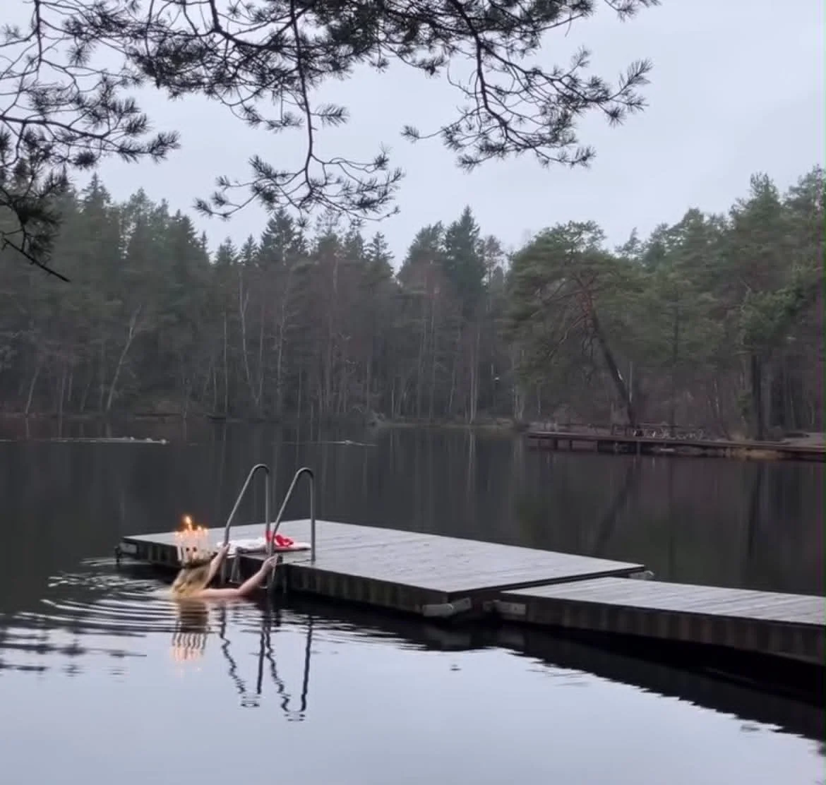 Person in the water holding onto a floating dock with candles on it, surrounded by calm lake and forested shoreline.