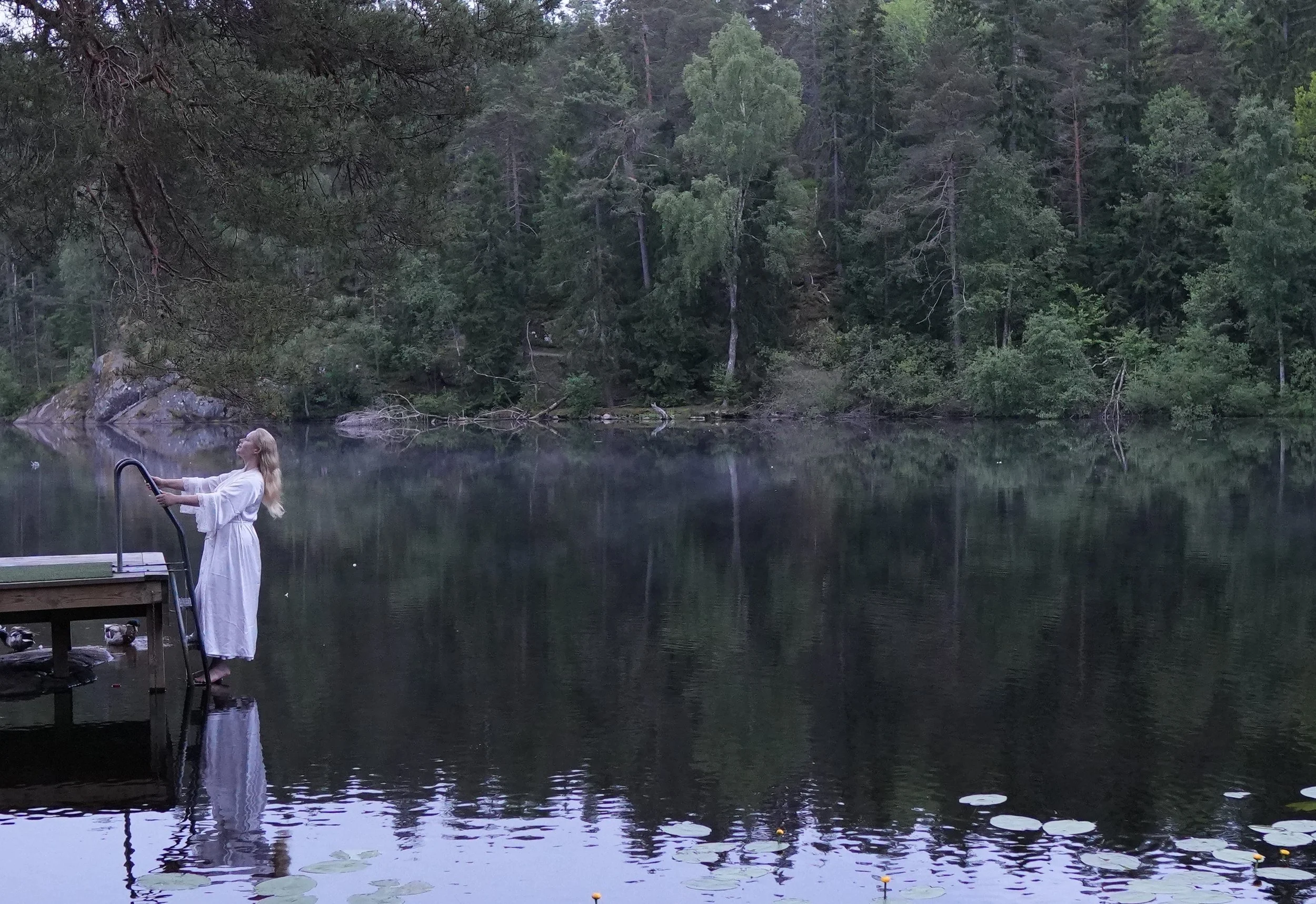 A woman in a white dress standing on a dock by a calm lake, surrounded by a dense forest of trees.