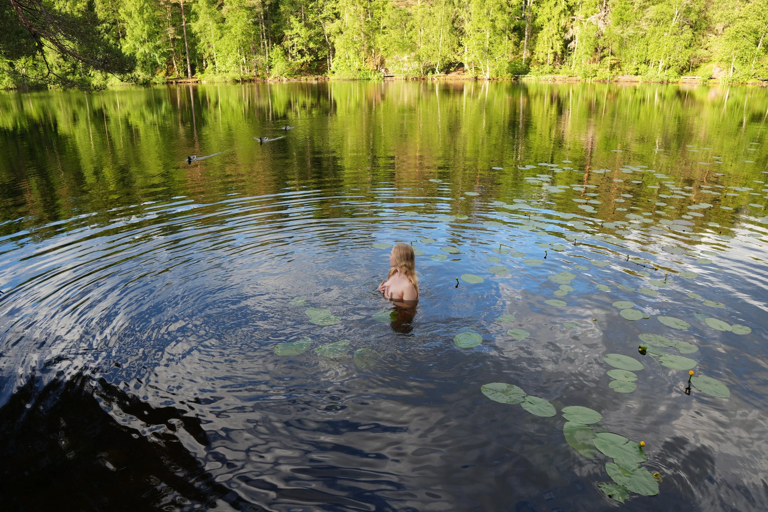 A woman with long blonde hair standing waist-deep in a calm pond with lily pads, surrounded by lush green trees on a sunny day.