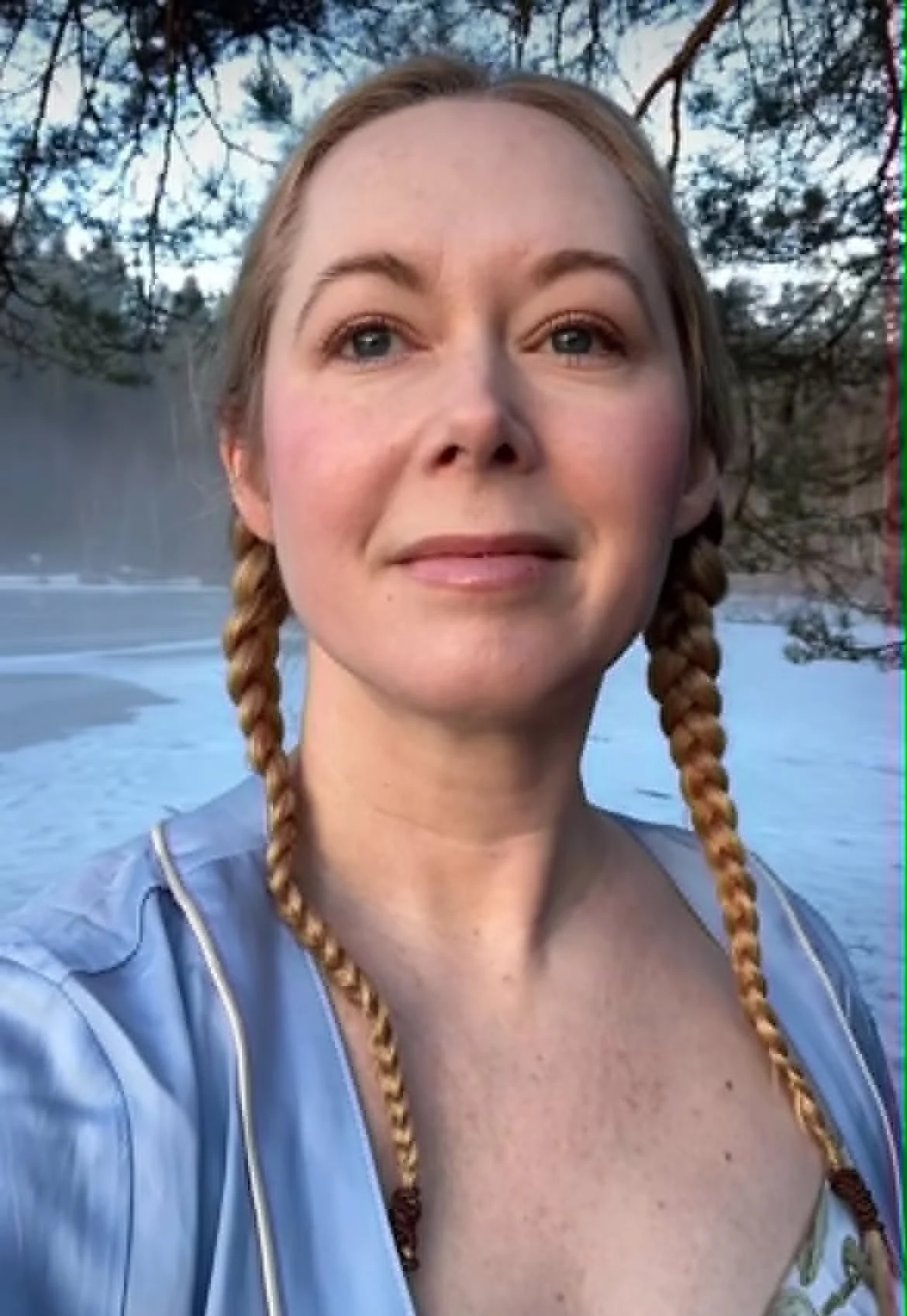 A woman with blonde hair in braids posing outdoors near trees with snow on the ground.
