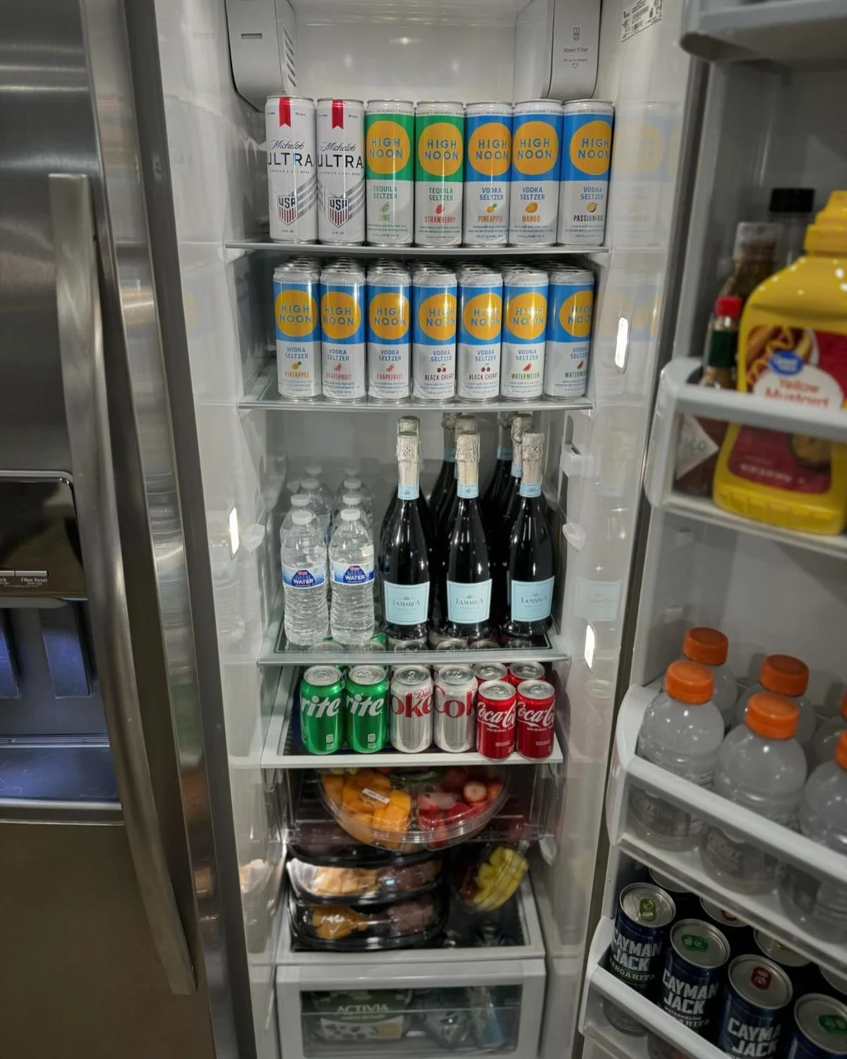 Inside a refrigerator stocked with beverages including energy drinks, flavored water, bottled water, bottles of wine, soda cans, and a bowl of fruit.