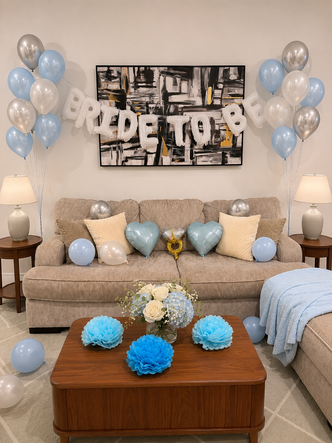 Living room decorated for a bridal shower with blue and silver balloons, a "BRIDE TO BE" banner, candles, a sofa with pillows, a floral center on a wooden table, and wall art.