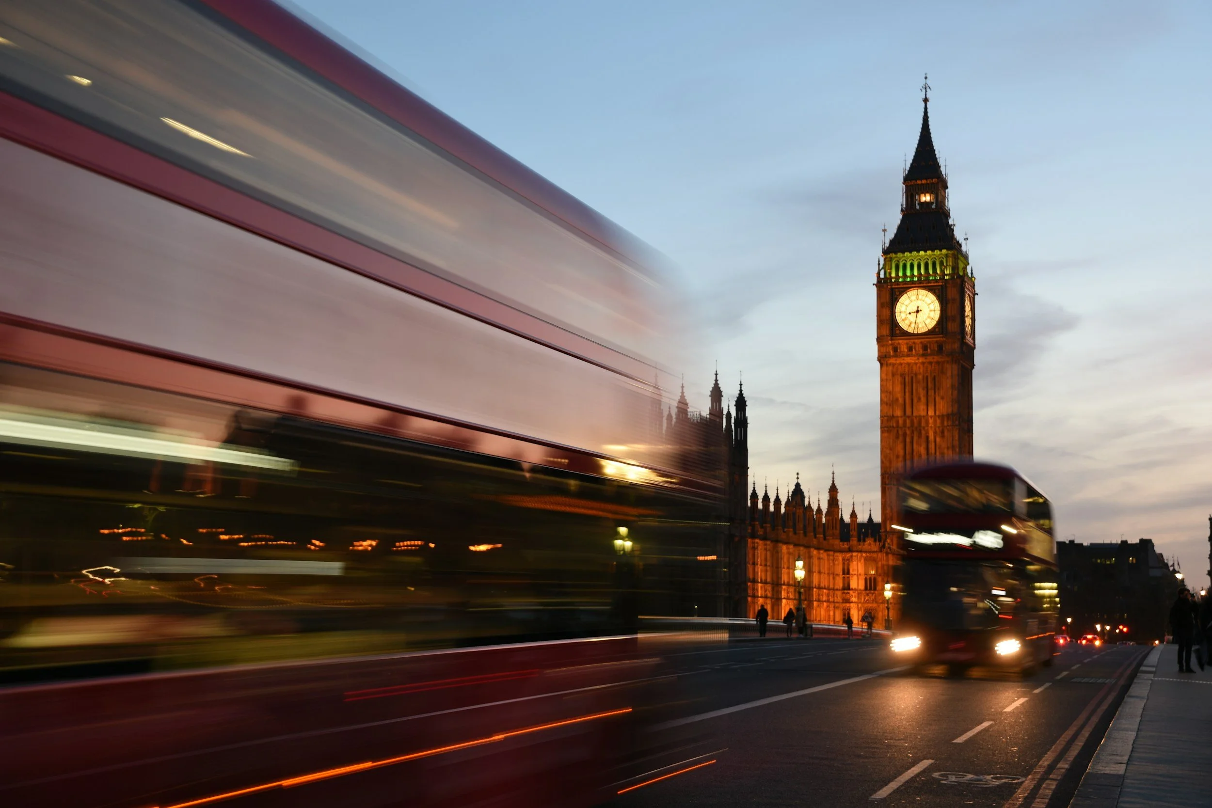 Blurred red double-decker bus and cars passing by Big Ben clock tower in London during dusk with a clear sky.