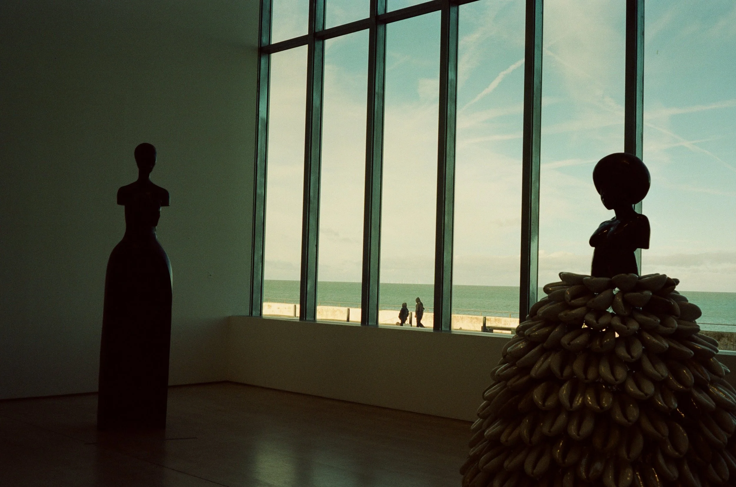 An indoor art installation of a sculpture made of numerous rocks stacked in a cone shape with a smooth, dark sphere on top, situated next to a large window with a view of the ocean and a blue sky with clouds. Two small human figures are seen outside 