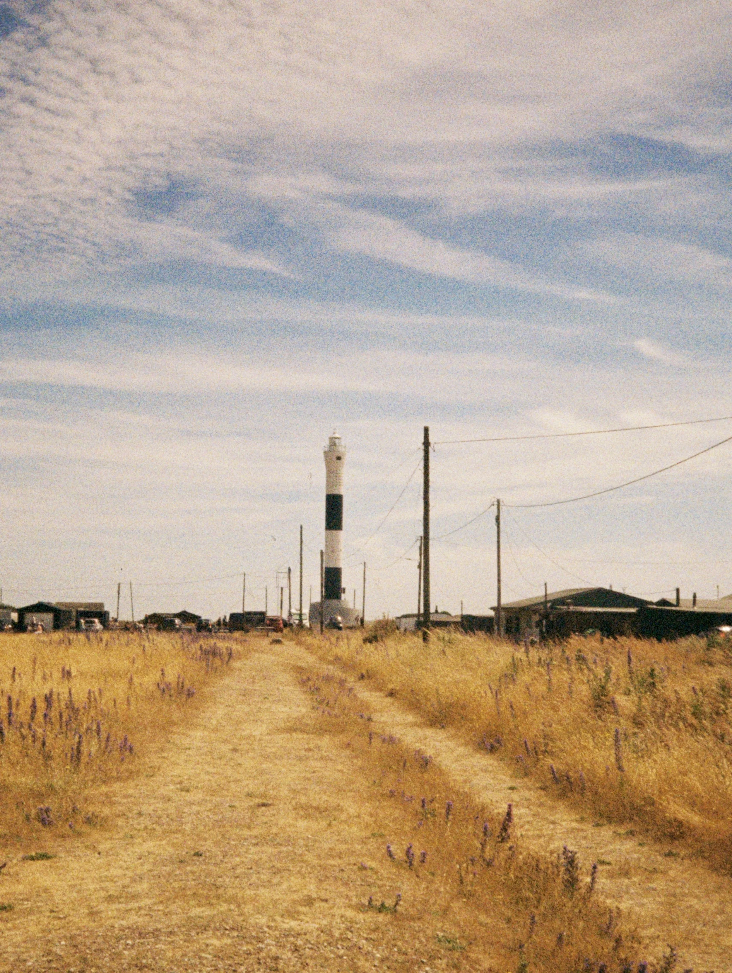 A lighthouse in a rural area with sparse buildings and utility poles, under a cloudy sky with various cloud formations.