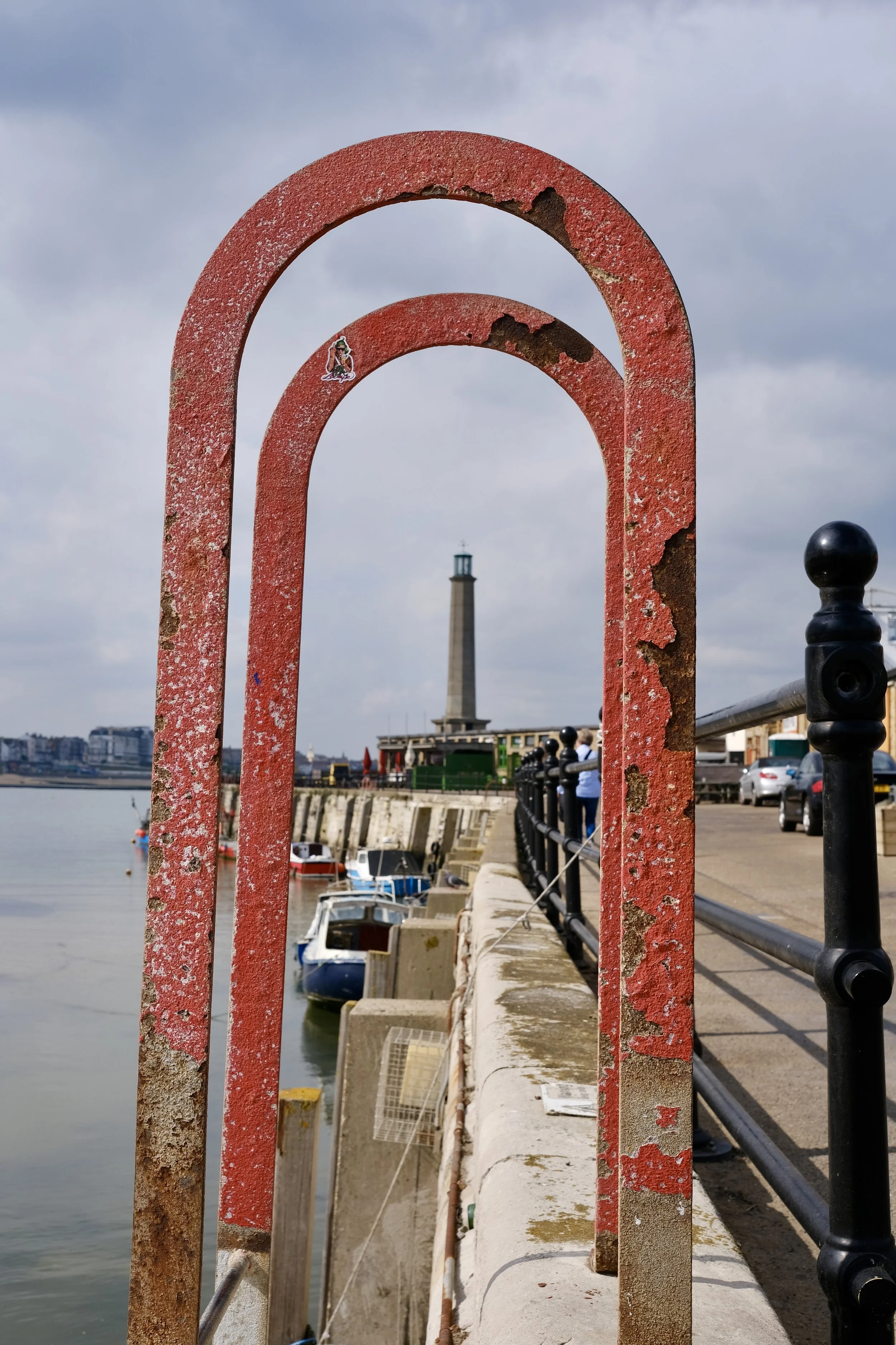 Rusty red metal arches along a waterfront promenade with boats docked below and a lighthouse in the distance under a cloudy sky.