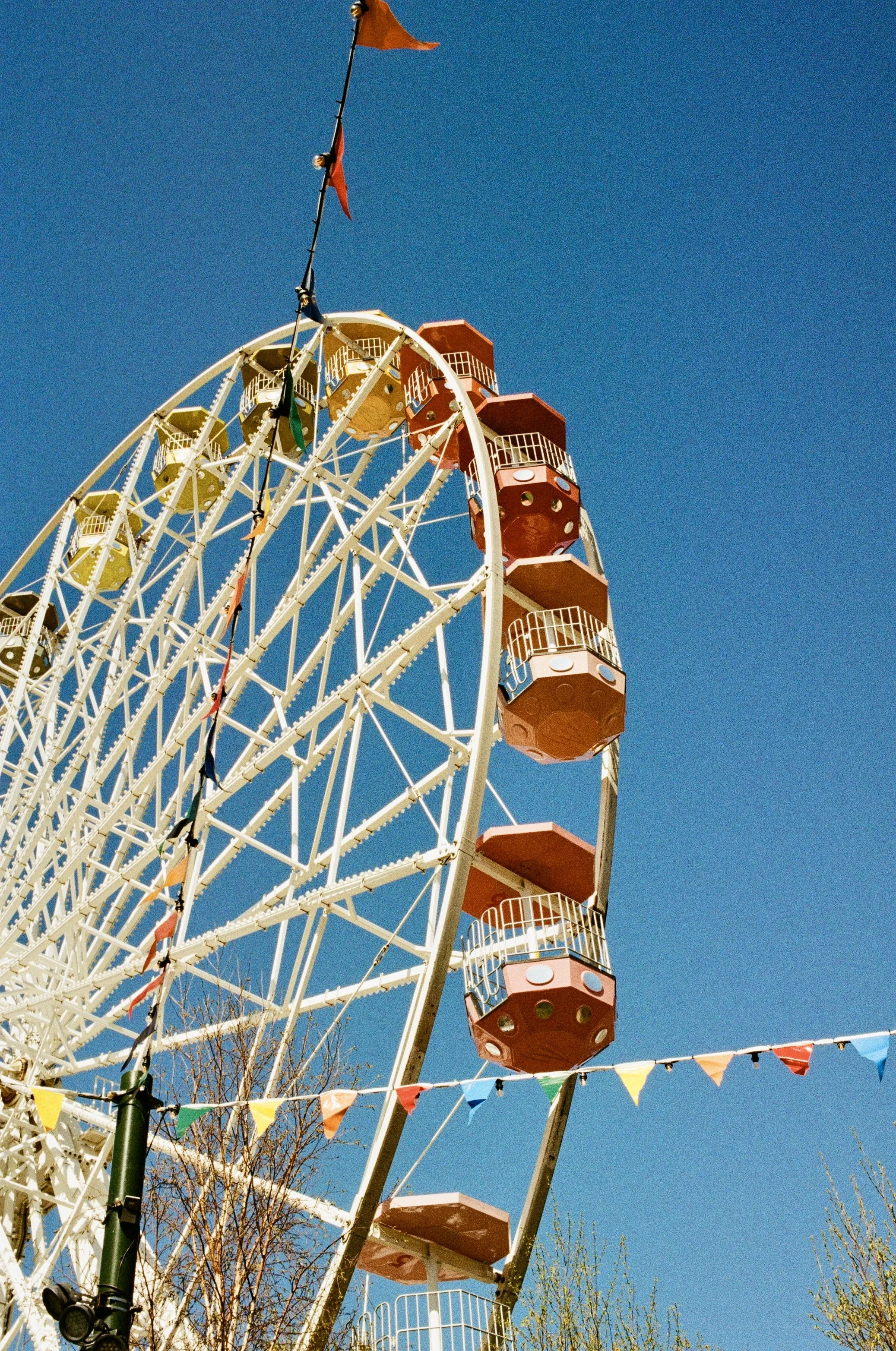 A large Ferris wheel at an amusement park with colorful flags strung in front. The Ferris wheel has red and yellow gondolas against a clear blue sky, with some leafless trees in the foreground.