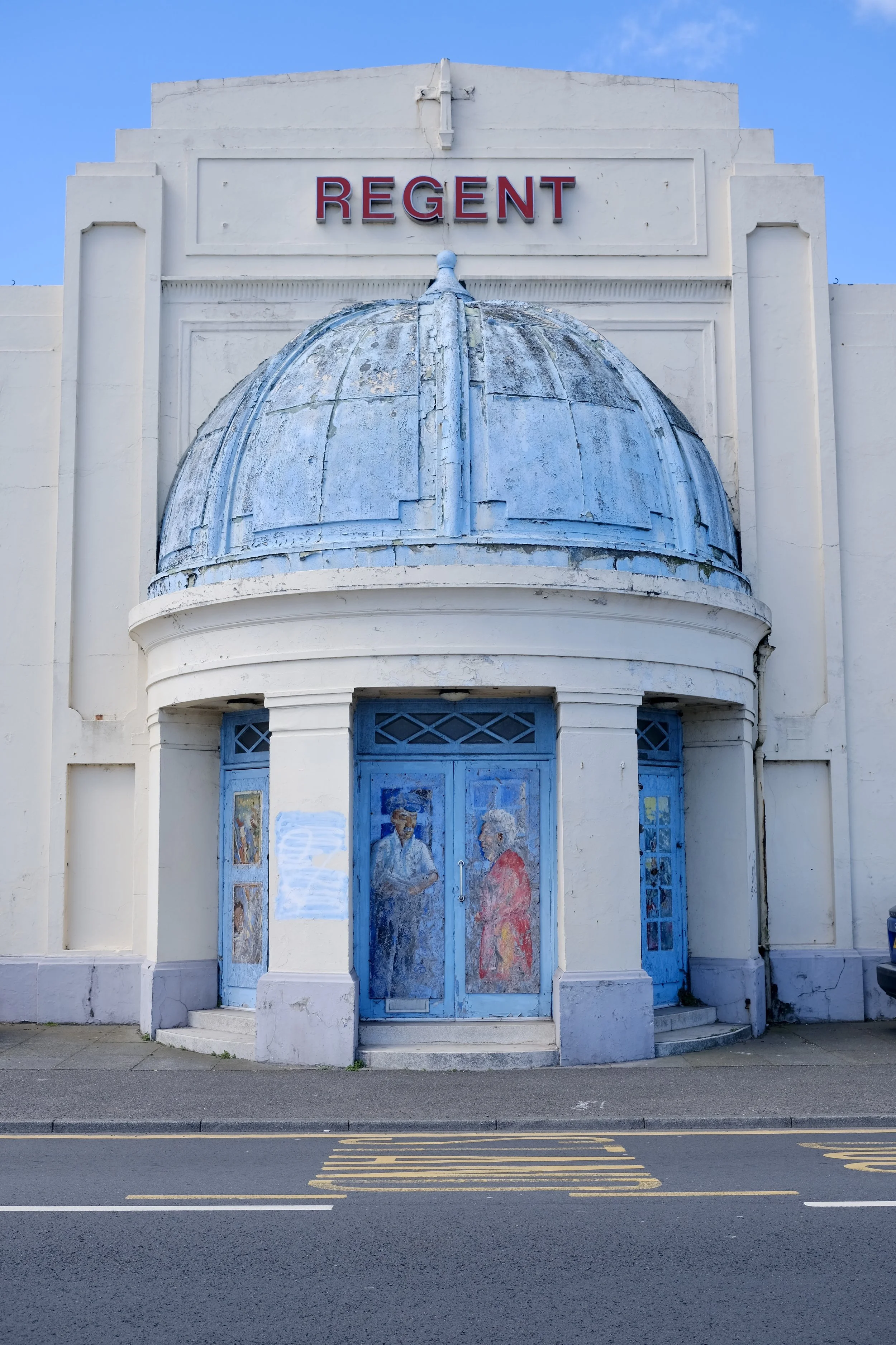 Front view of an old, white art deco building with a dome and blue doors, with a sign that says 'REGENT' at the top. The building's paint is chipped, and there are murals on the doors. In front is a street with yellow parking lines.