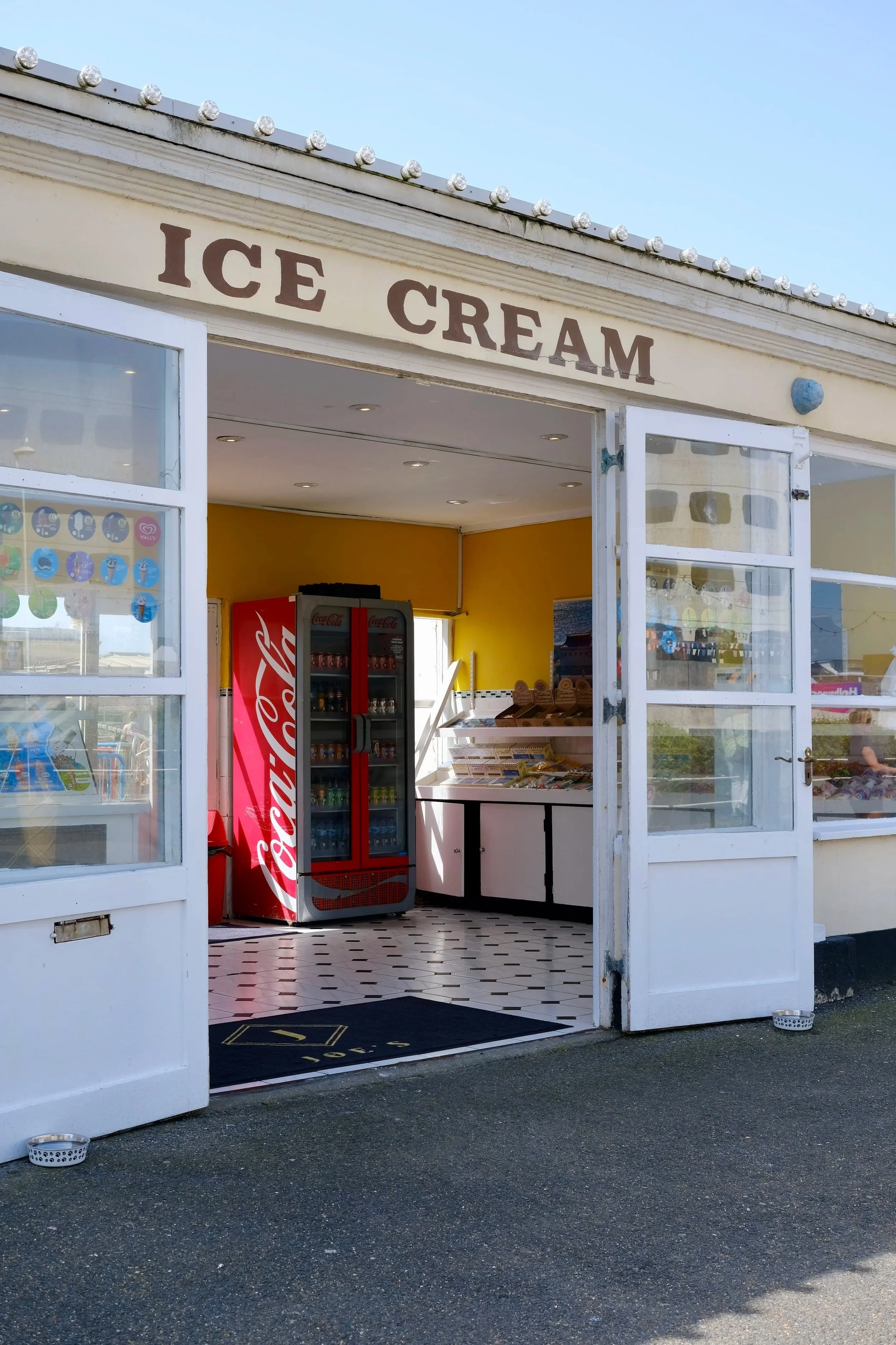 A small ice cream shop with open white doors, a Coca-Cola refrigerator, and a yellow interior wall. The shop has a black and white checkered floor and outside there are two small round pet bowls on the pavement.