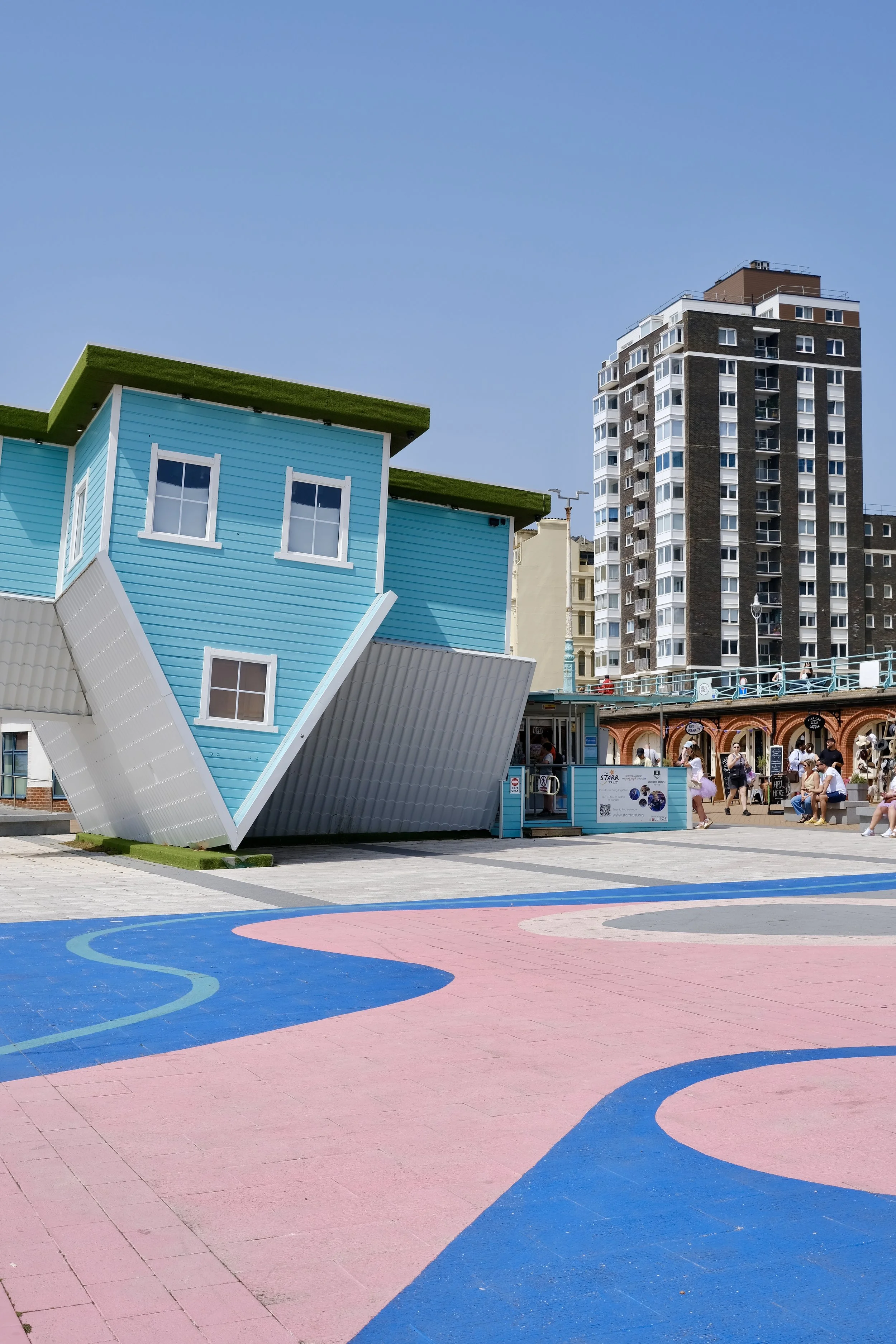 Colorful outdoor basketball court with pink and blue surface, modern blue building with white windows, and high-rise apartment building in the background under a clear blue sky, with people walking nearby.