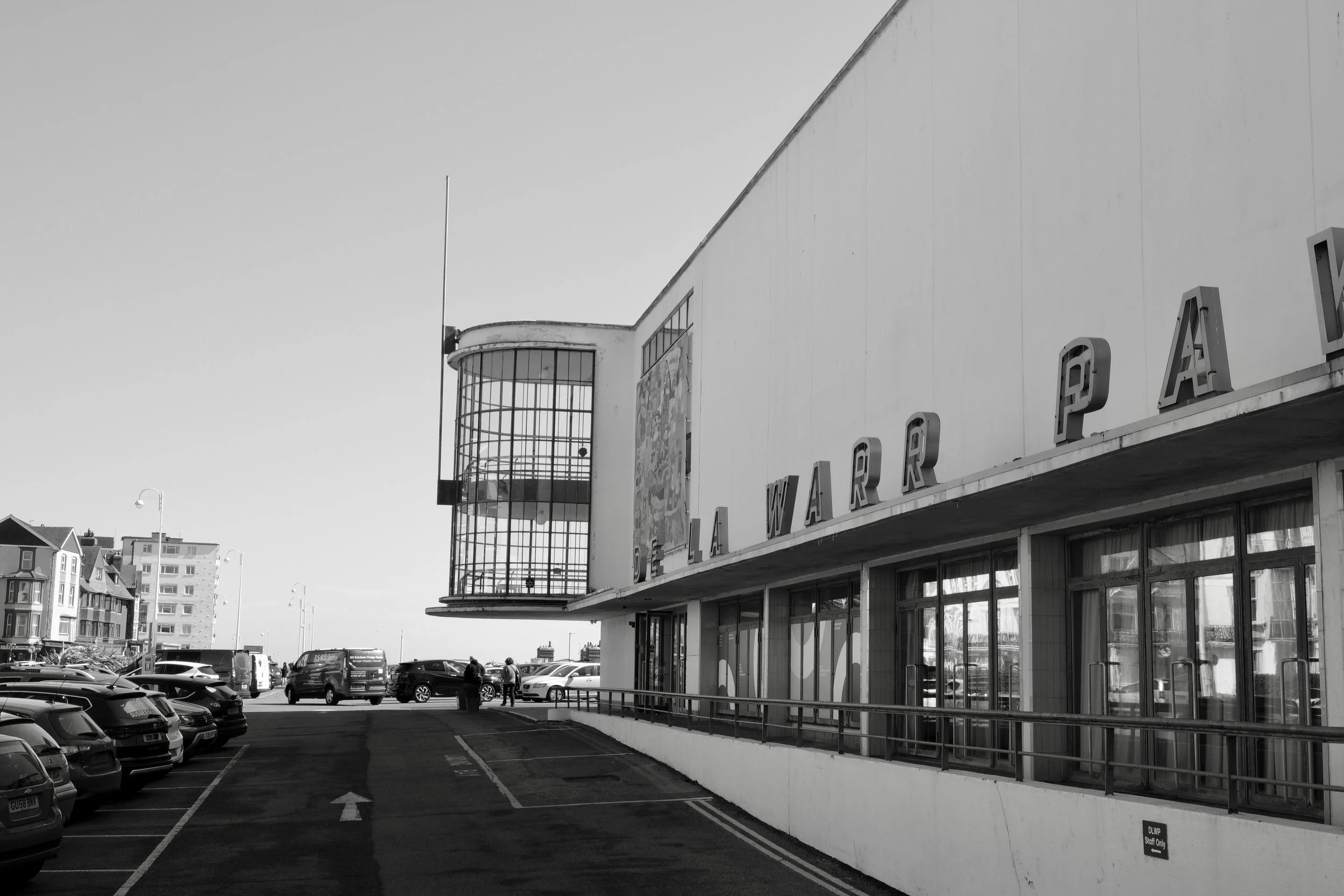A black and white photo of a parking lot in front of a building with a sign that reads 'Galeria.' The building has large glass windows and a curved section at the top. Several cars are parked, and a few people are walking near the entrance.