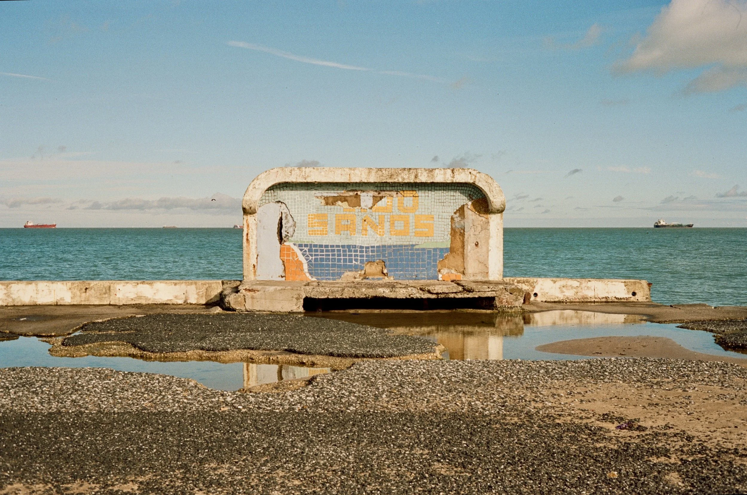 A weathered, broken metal sign with the words 'NO TAPS' in faded yellow paint, situated on a concrete and pebbled surface near the ocean with ships in the distance under a partly cloudy sky.