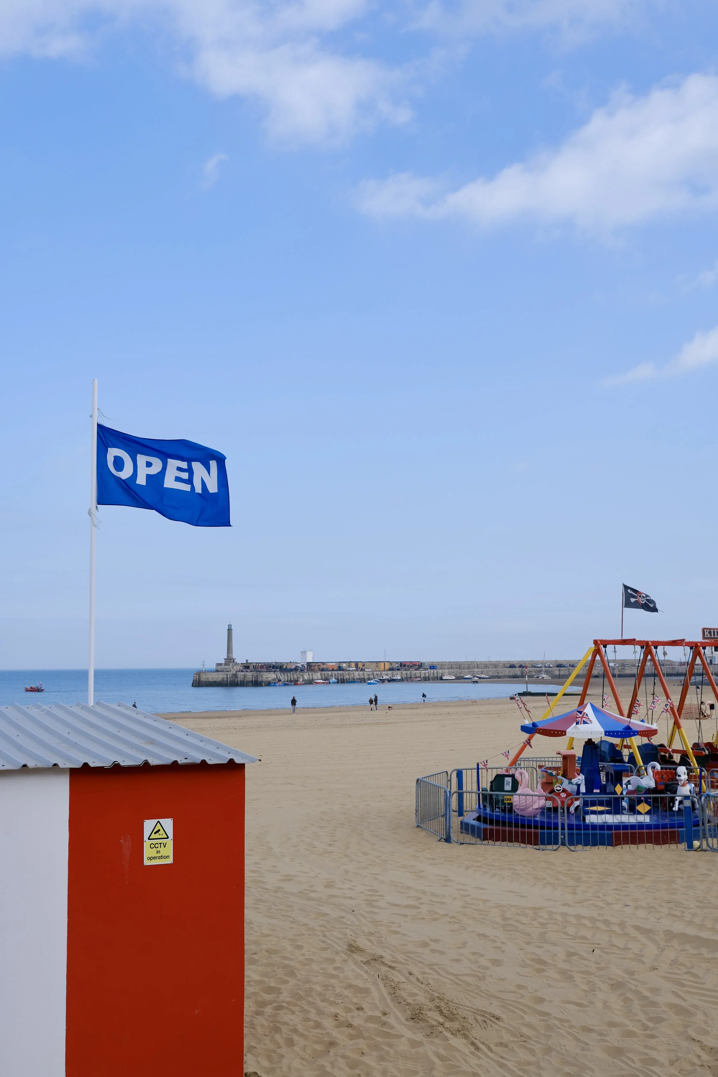 A beach with a blue flag that says 'OPEN', a red and white small shack with a CCTV sign, and a playground with a swing nearby. In the background, there are people walking, a pier, and a lighthouse, with a partly cloudy sky.