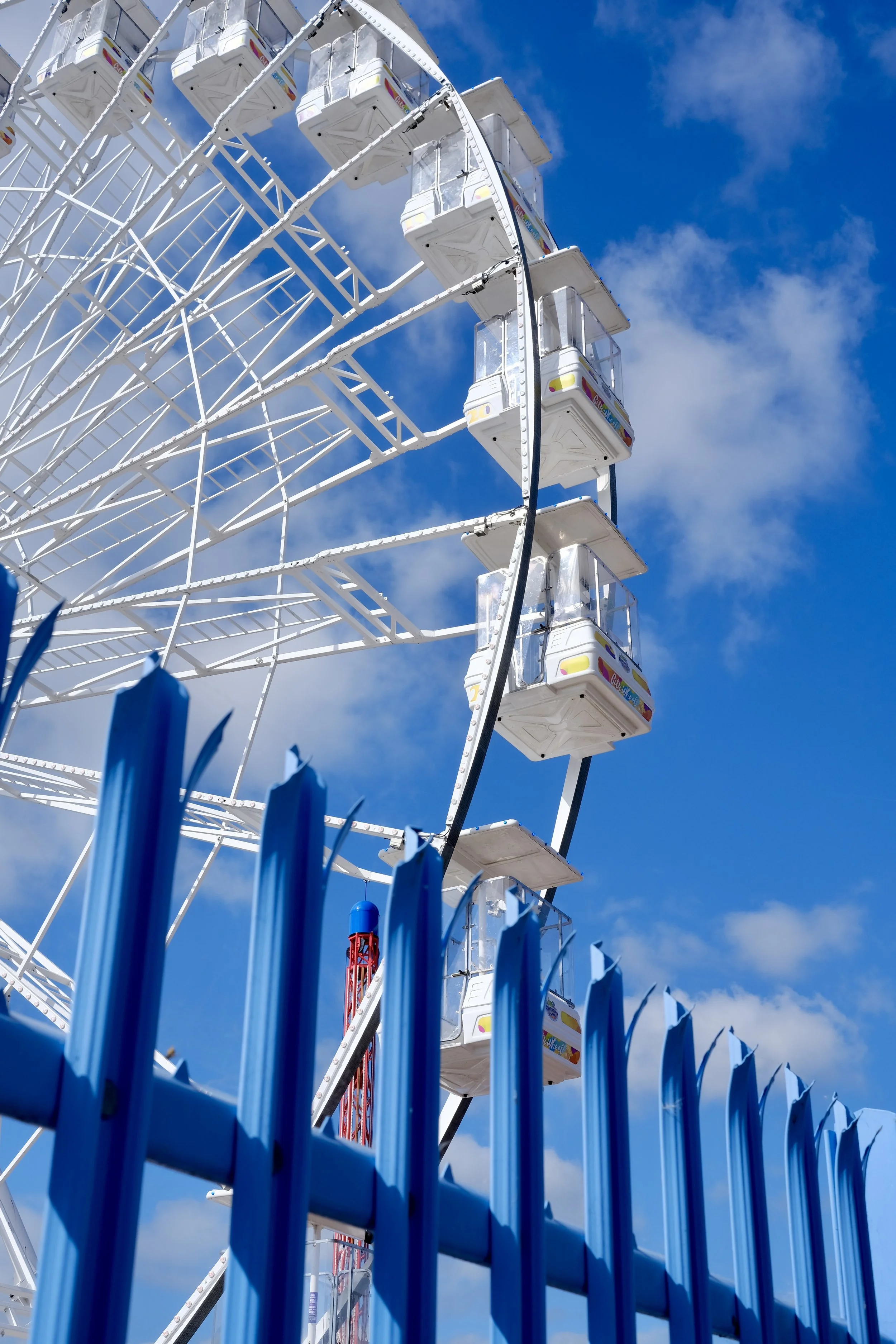 A Ferris wheel with enclosed cabins behind a blue metal fence, set against a blue sky with some clouds.