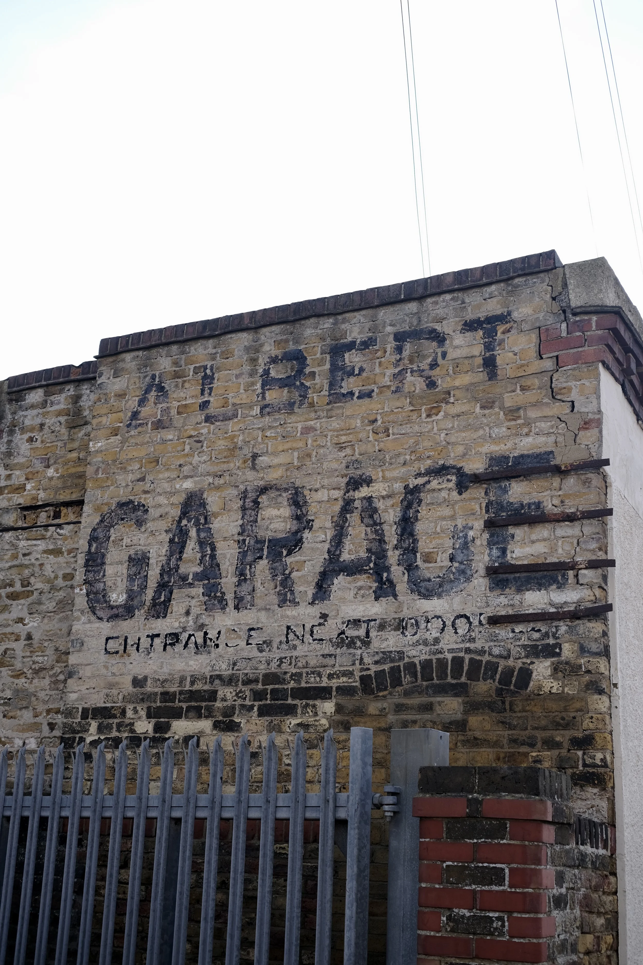 A brick wall with faded black painted letters reading 'V G P.' below, it says 'Entrance Next Door.' A metal gate with spikes and brick columns is in front of the wall.