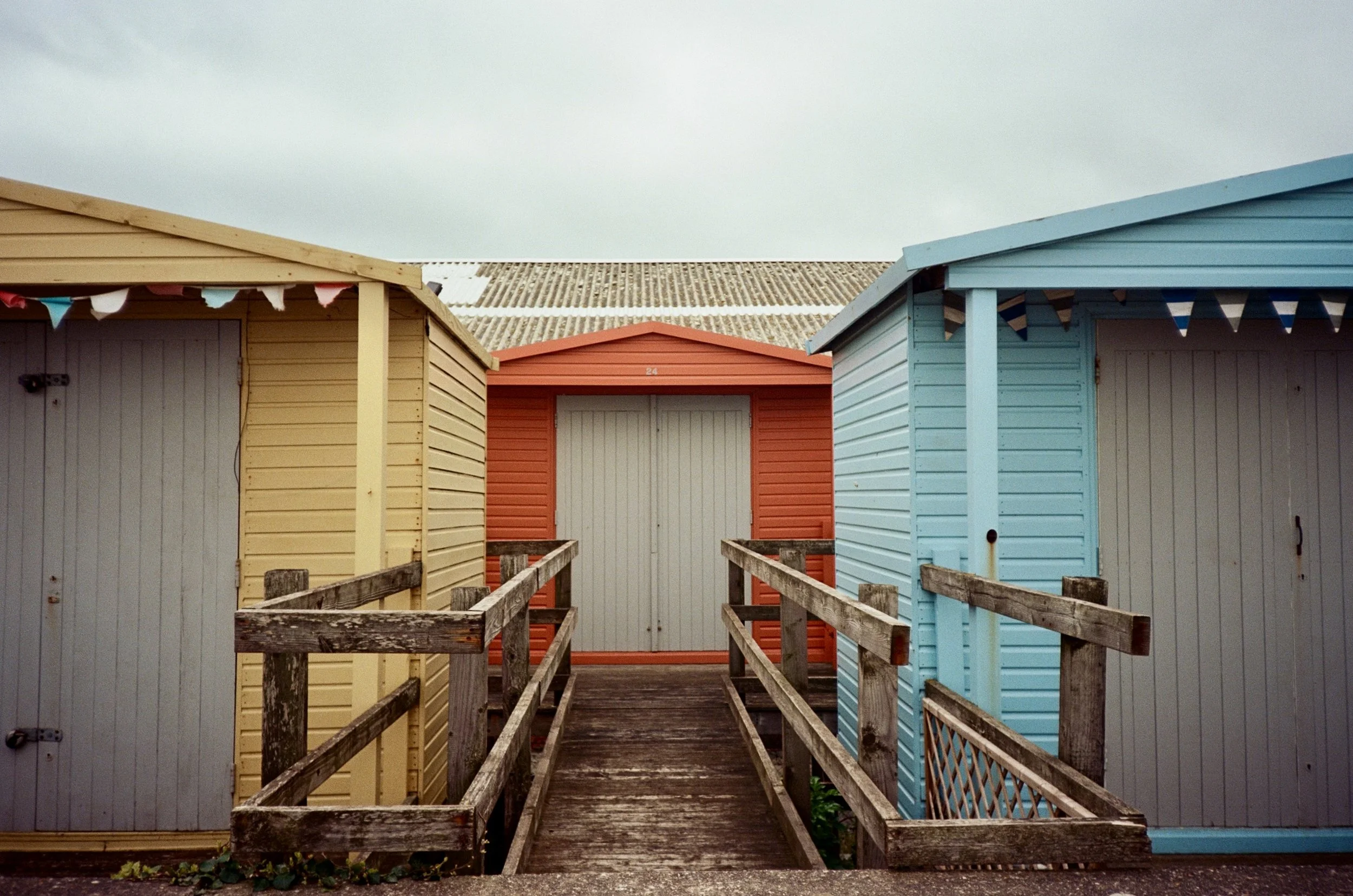 Colorful beach huts painted in yellow, red, and blue, connected by a wooden walkway with railings, under an overcast sky.