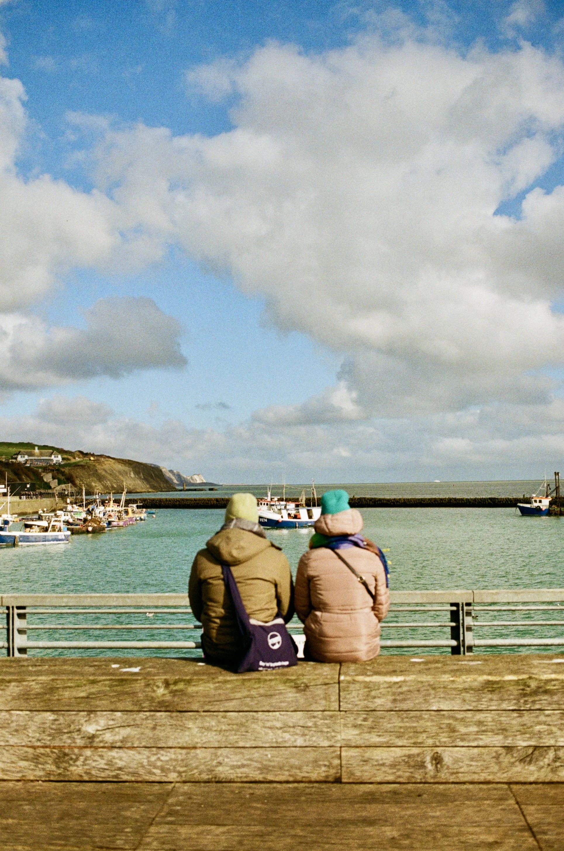 Two people sitting on a wooden bench at a marina, overlooking boats docked in the water with a coastal landscape and cloudy sky in the background.