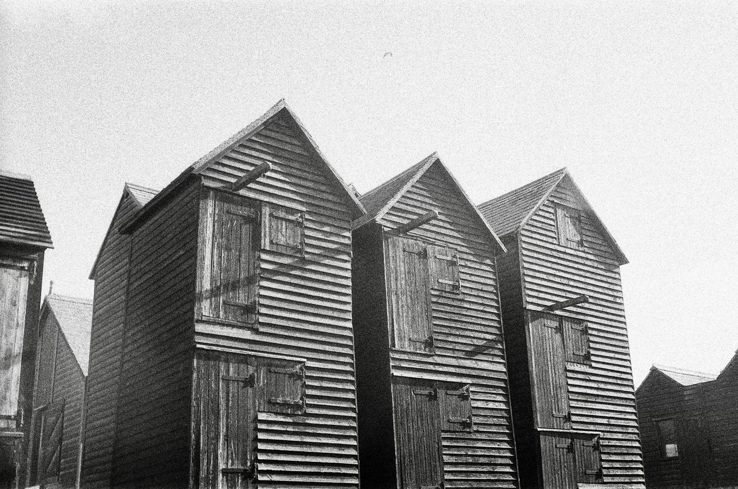Black and white photo of old wooden houses with steeply pitched roofs and boarded-up windows, with a cloudy sky in the background.