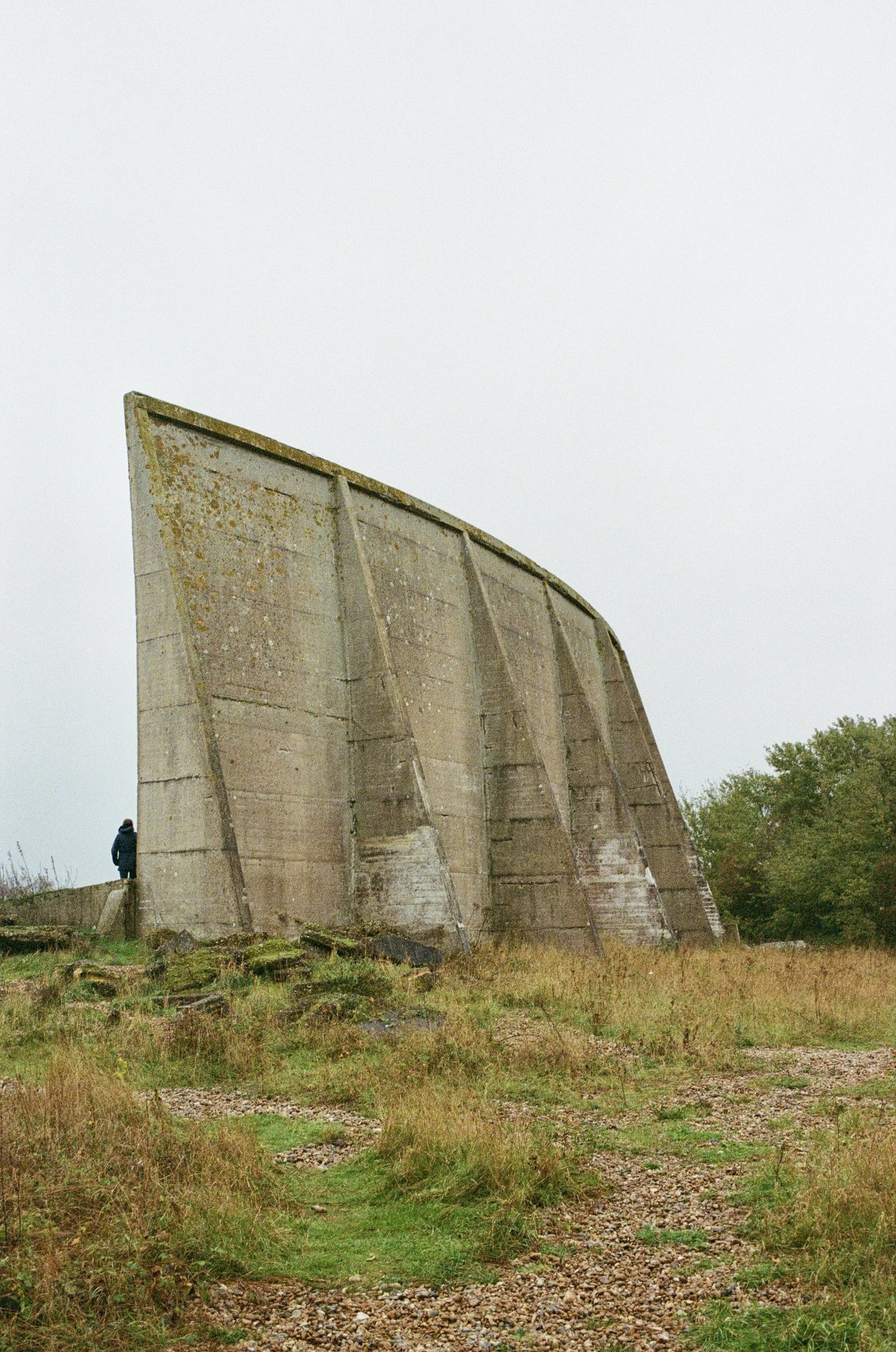 A large, weathered concrete structure with a curved top, rusted and moss-covered, set against a cloudy sky. A person in dark clothing stands near the base of the structure, amidst grassy and rocky terrain.