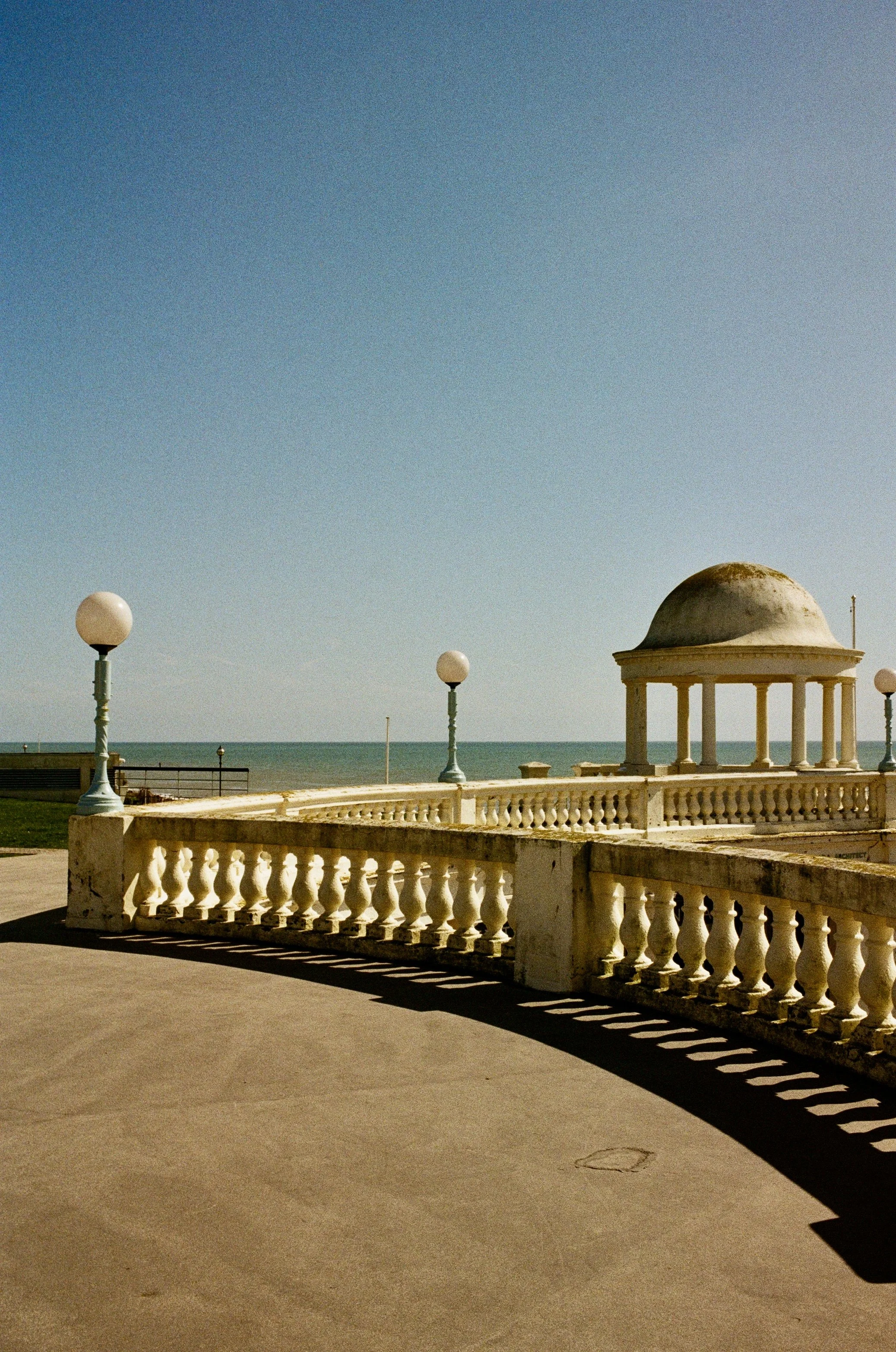 Beachside promenade with white balustrade, several lamp posts, and a domed pavilion, overlooking the ocean under a clear blue sky.