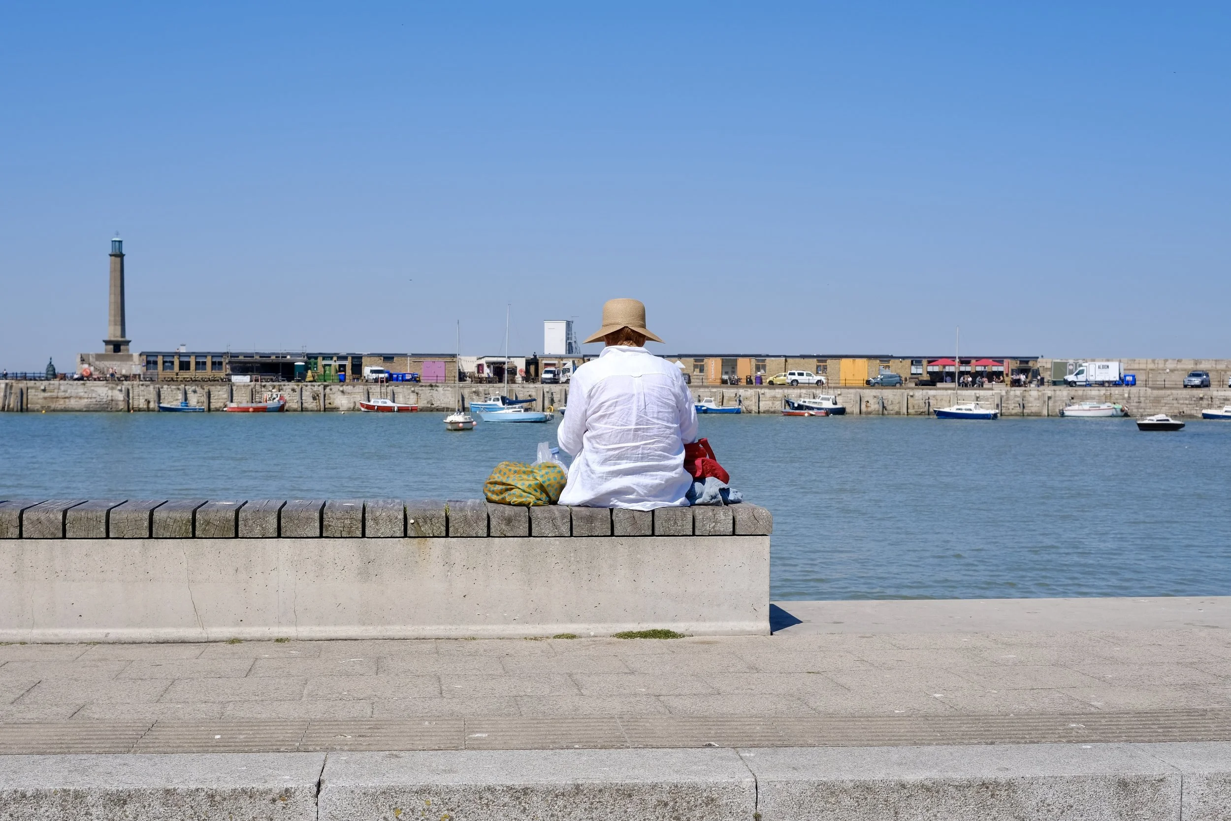 A person sitting on a stone bench by the water, facing away, wearing a wide-brimmed hat and white shirt, with boats and a harbor in the background under a blue sky.