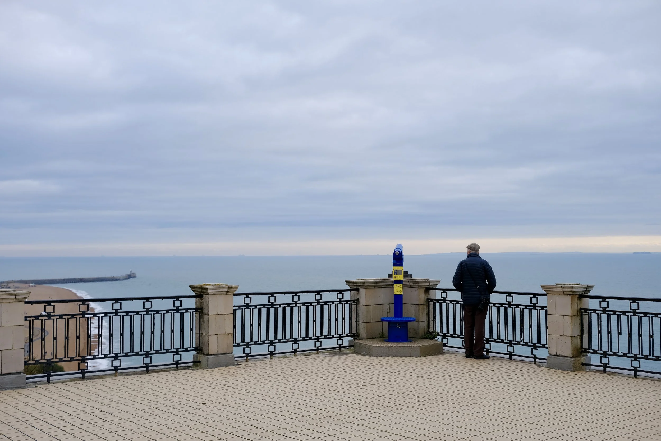 A man standing on a balcony overlooking the ocean under a cloudy sky, with a blue telescope-like viewing device nearby.