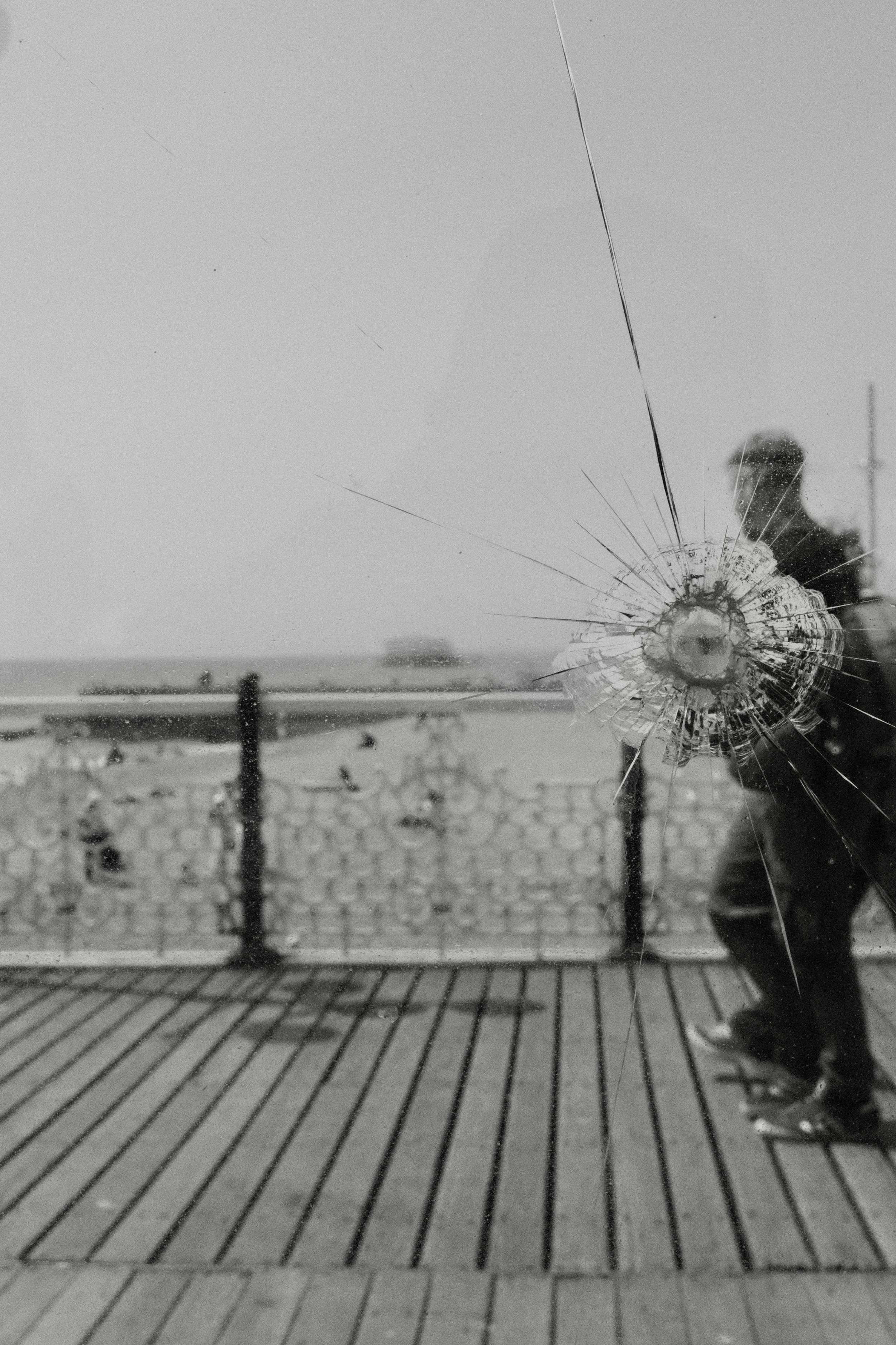 A shattered glass window with a large hole and cracks radiating outward, showing a person standing outside on a windy day with snow on the ground and a fence in the background.