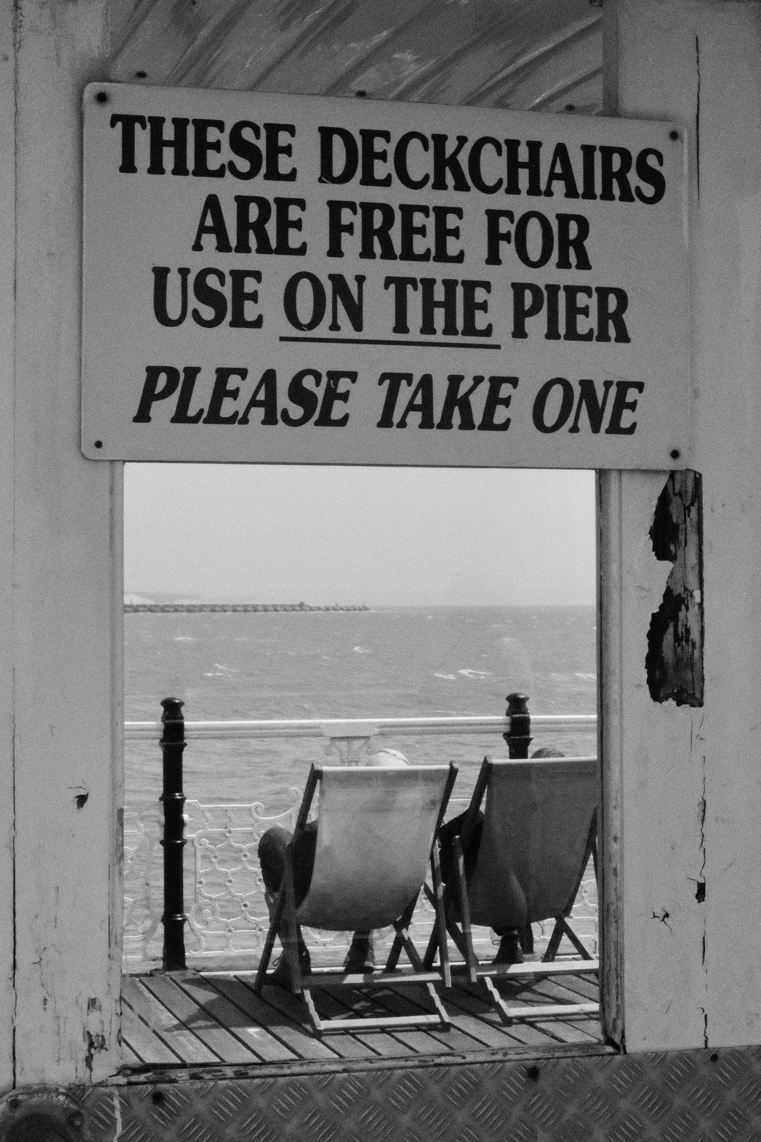 A sign that reads, 'These deckchairs are free for use on the pier, please take one', seen through a wooden doorway. In the background, there are deckchairs facing the water, with a view of the sea.