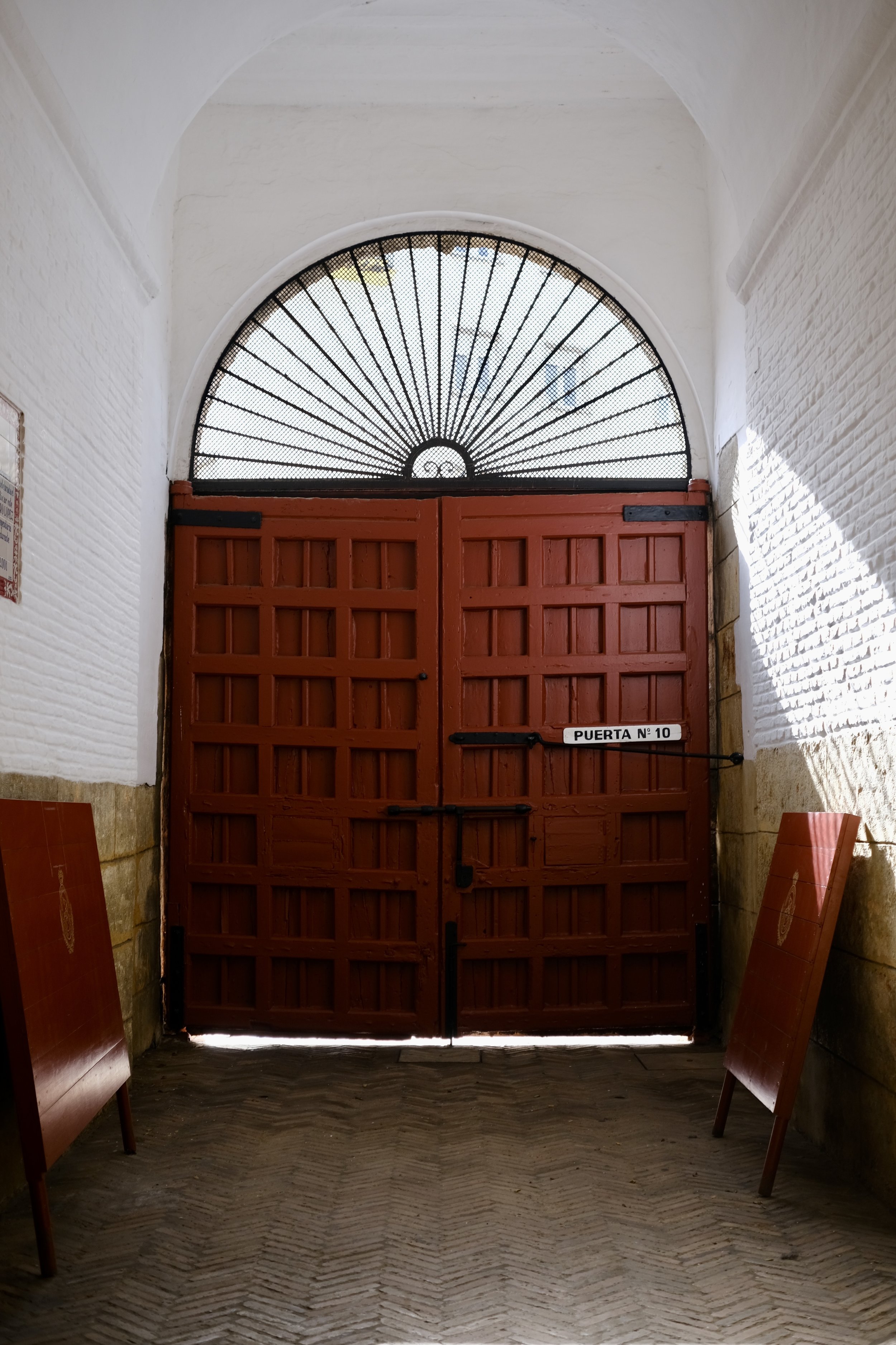 A red wooden gate with black hinges and a sign that reads 'PUERTA N° 10'. The gate has a semi-circular metal grate with radiating lines above it, allowing light to filter through. The surrounding walls are made of white-painted brick and the floor ha