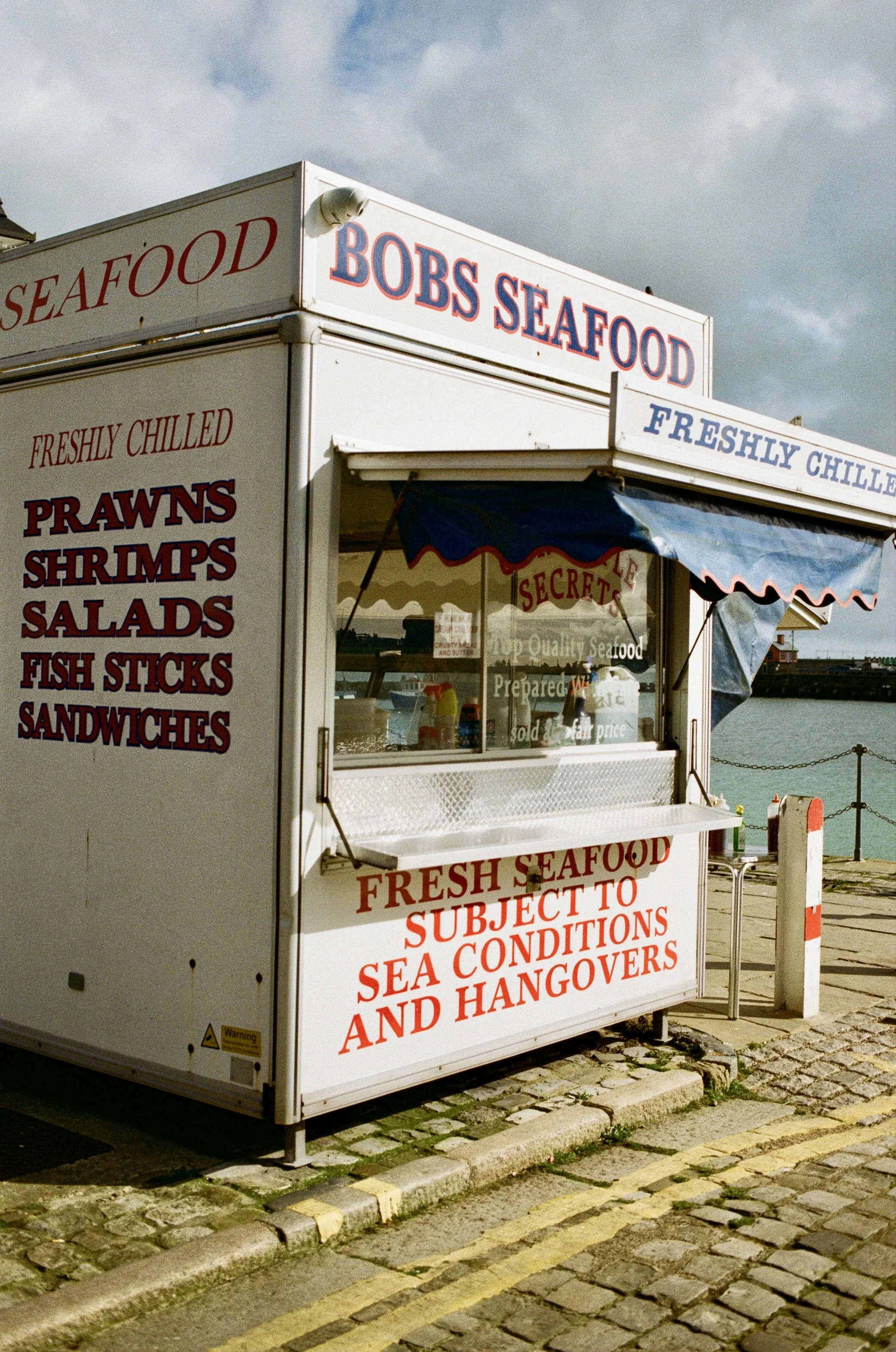 A white seafood stand with signs reading "Bobs Seafood," "Freshly Chilled," and a menu listing prawns, shrimps, salads, fish sticks, and sandwiches, situated on a cobblestone street near water under a cloudy sky.