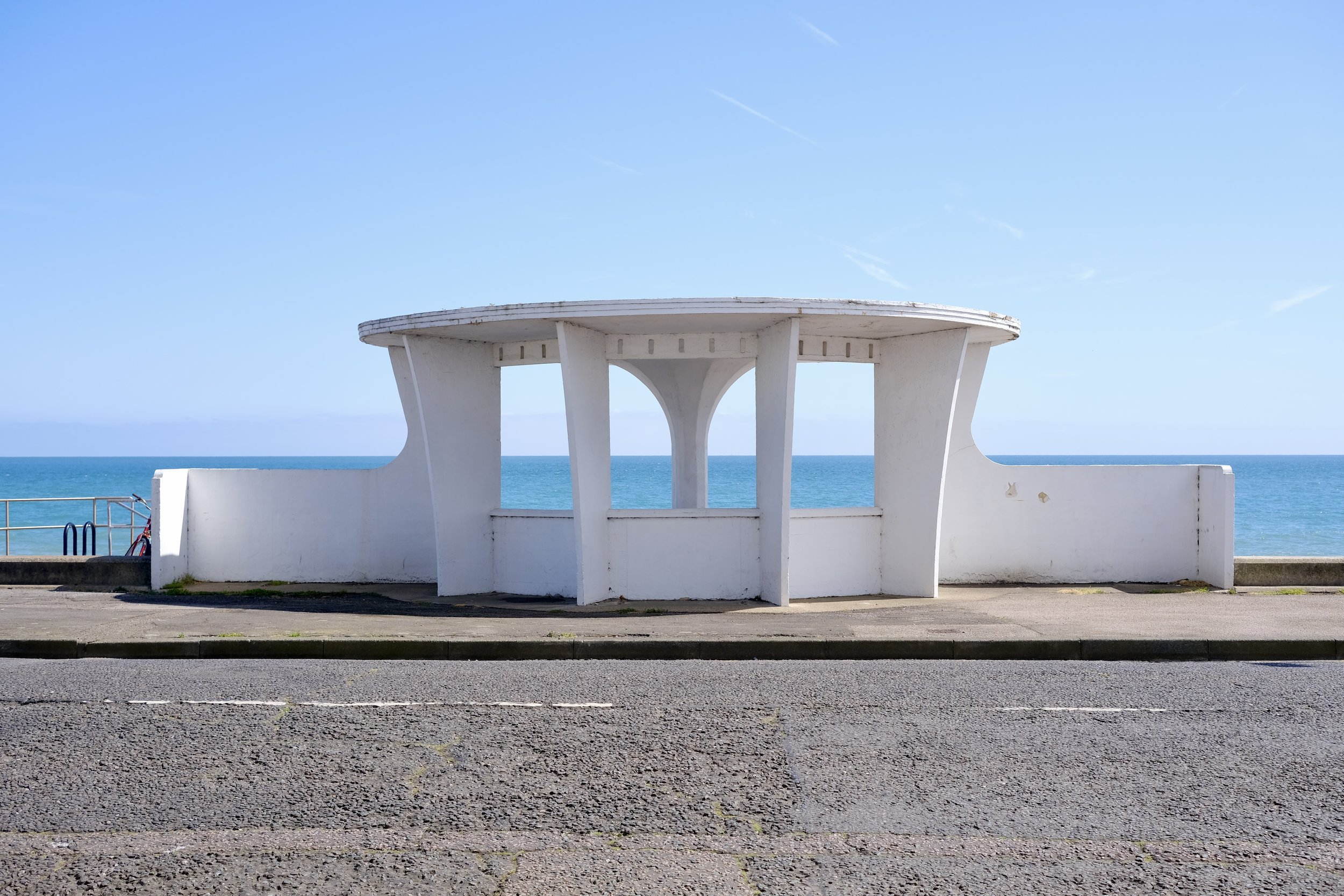 White modern pavilion structure overlooking the ocean, set against a clear blue sky with a few wispy clouds.