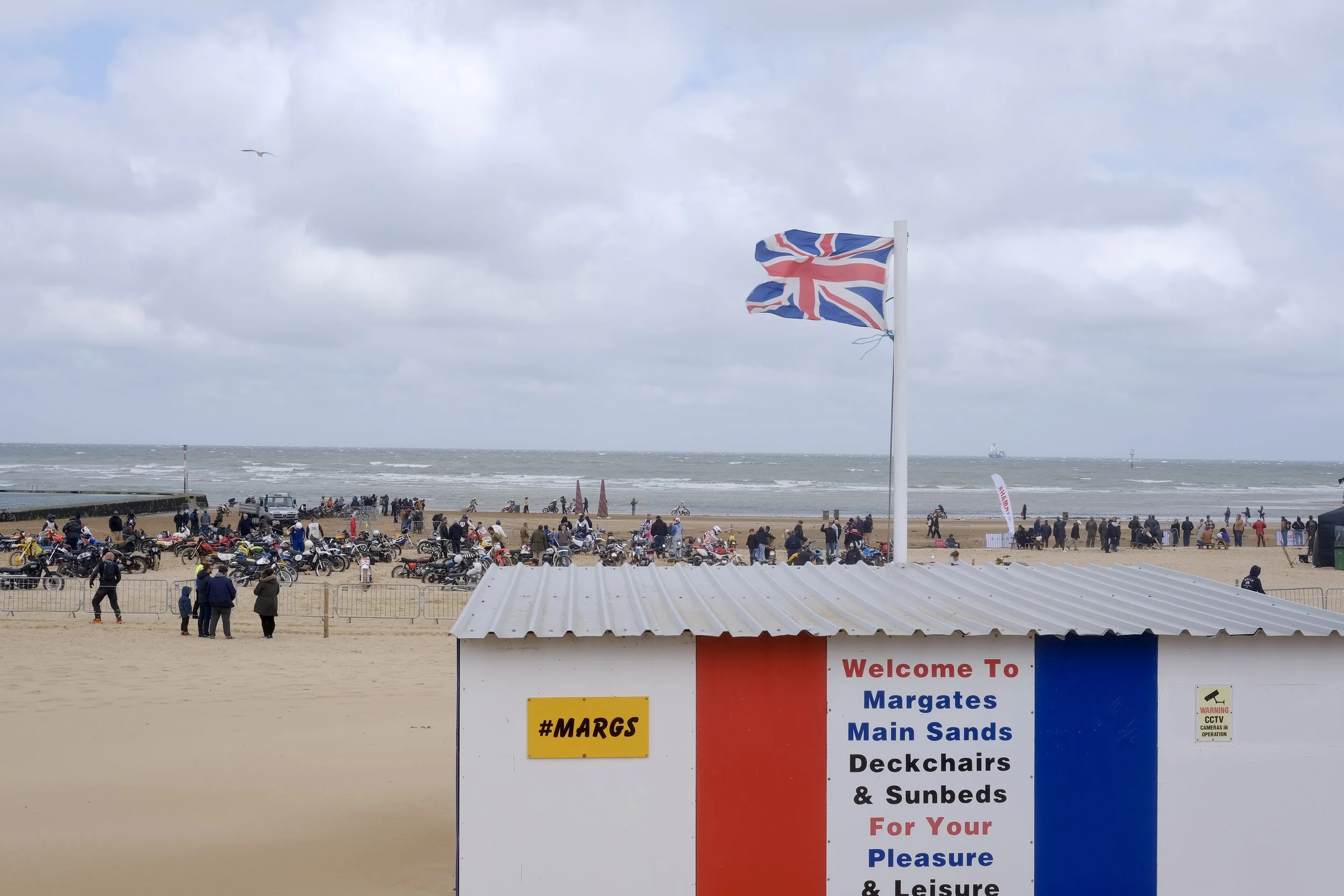 Beach scene with a large British flag on a pole, cloudy sky, people walking and gathering, numerous motorcycles parked on the sand, and a sign welcoming visitors to Margate with details about disabilities and sunbeds available for pleasure and leisur