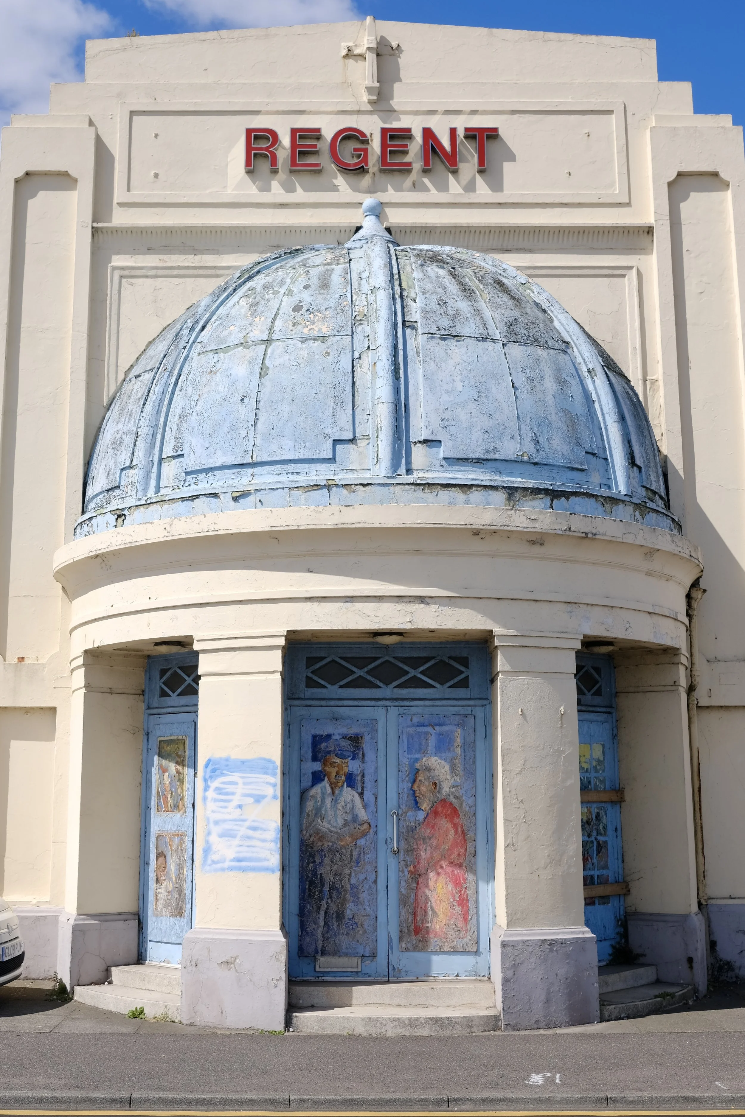 Facade of a building with a sign reading 'Regent' in red letters, featuring an art deco style, with a rounded, domed awning above the entrance, and murals or painted panels on the doors.