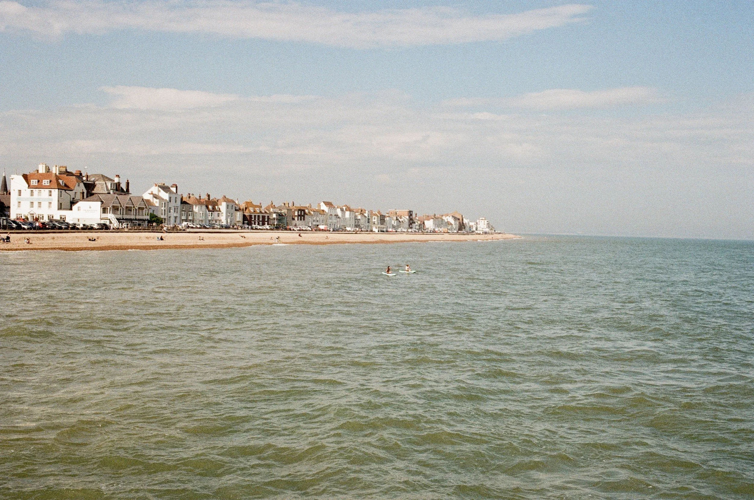 A coastal scene with a sandy beach and colorful buildings along the shoreline. The water has gentle waves, and there are a few people paddleboarding in the distance. The sky is partly cloudy.