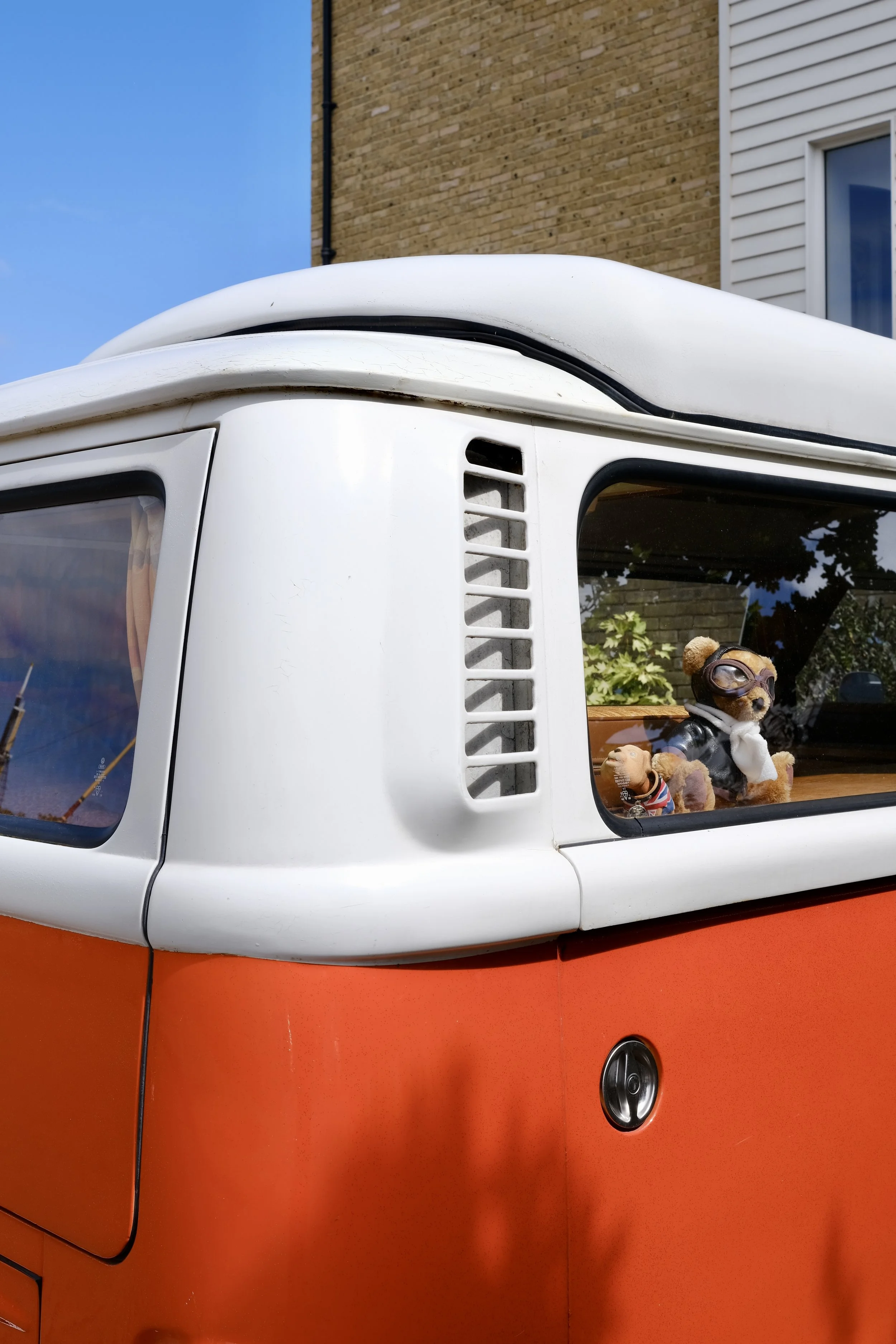 Close-up of a vintage orange and white vehicle, showing part of the side and a window with two teddy bears dressed in costumes inside.